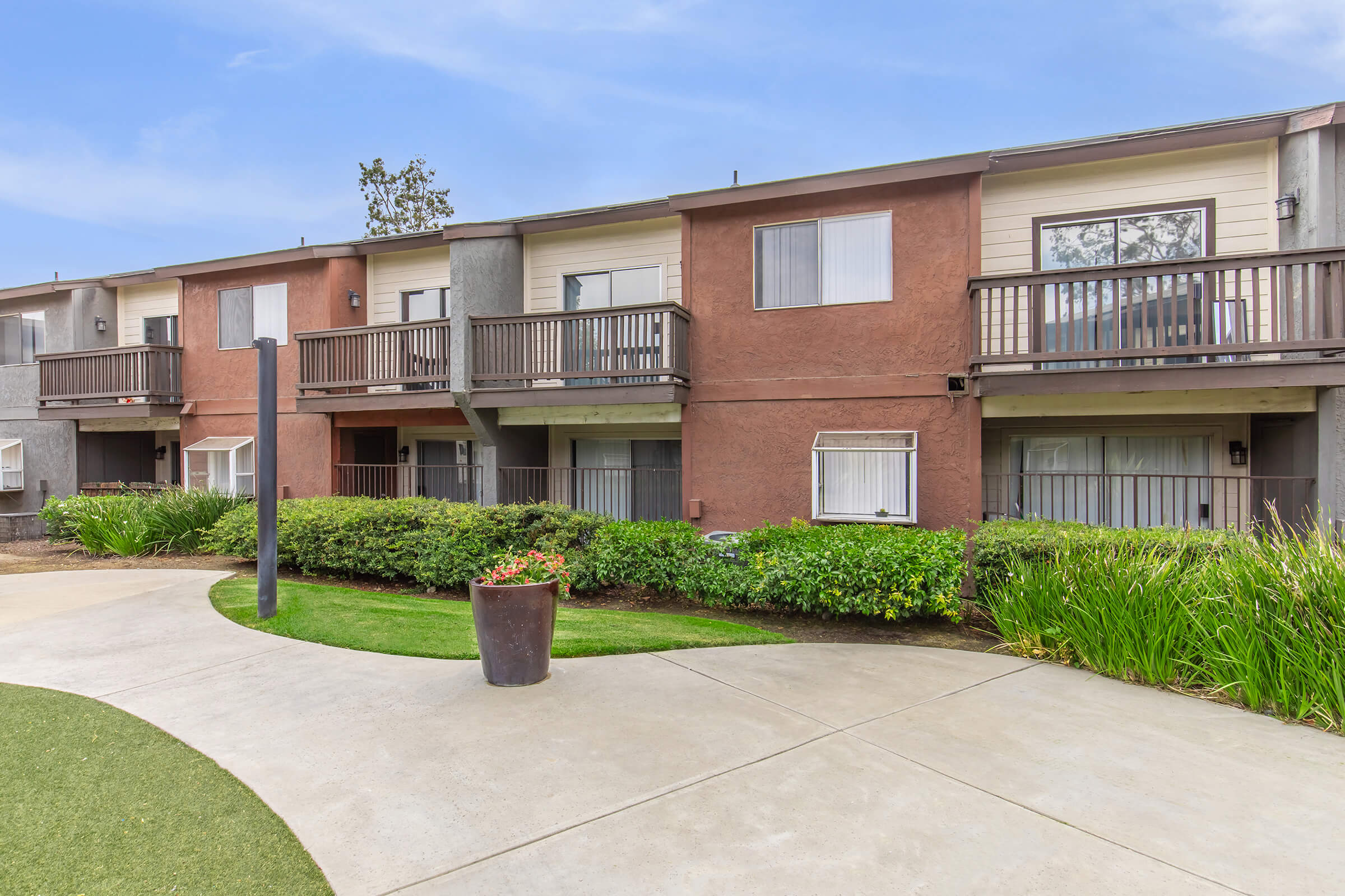 A landscaped courtyard featuring two-story buildings with balconies. The pathways are paved, leading through green grass and plants. A flower pot is visible near the pathway, with a clear blue sky in the background. The setting appears to be a residential area or apartment complex.