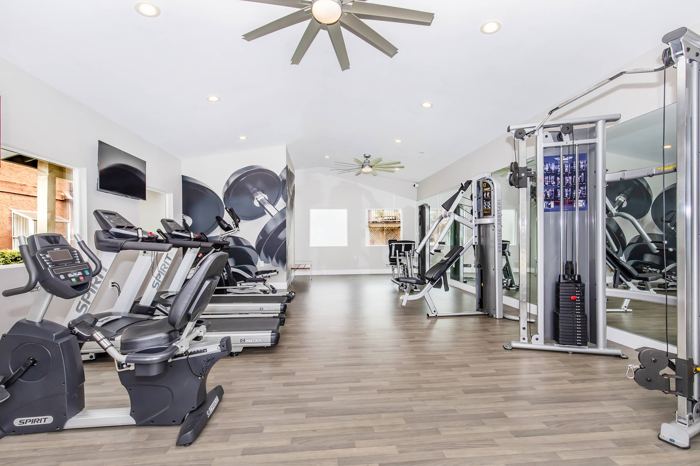 A bright and modern gym featuring various exercise equipment, including treadmills, stationary bikes, and a multi-gym setup. The space has large windows allowing natural light, a ceiling fan, and contemporary decor, creating an inviting workout environment.
