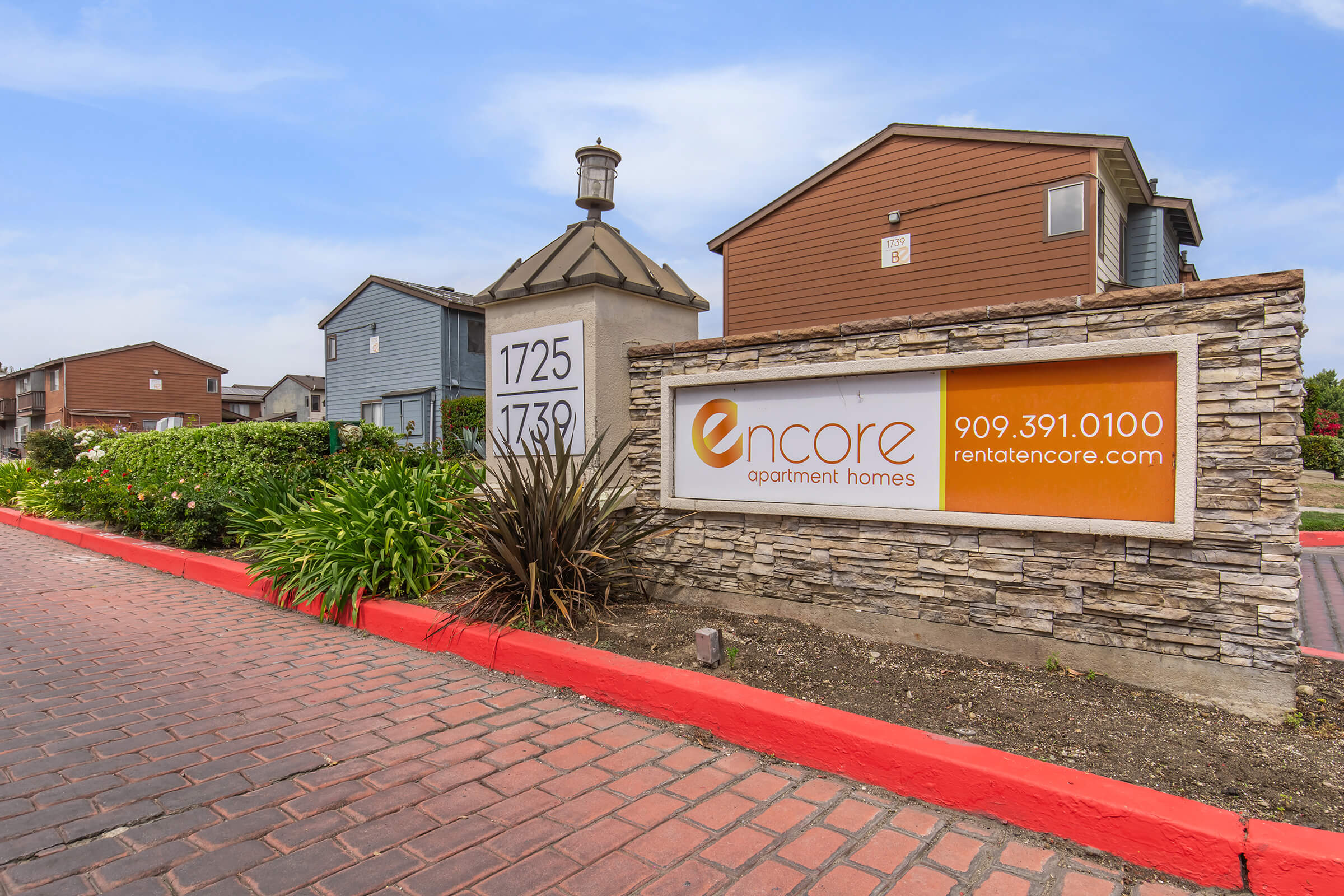 Sign for Encore Apartment Homes, featuring the address numbers 1725 and 1739. The sign is set against a landscaped area with colorful plants, and there's a clear blue sky above. The contact number 909.391.0100 and website rentencore.com are visible. The entrance is bordered by red paving stones.