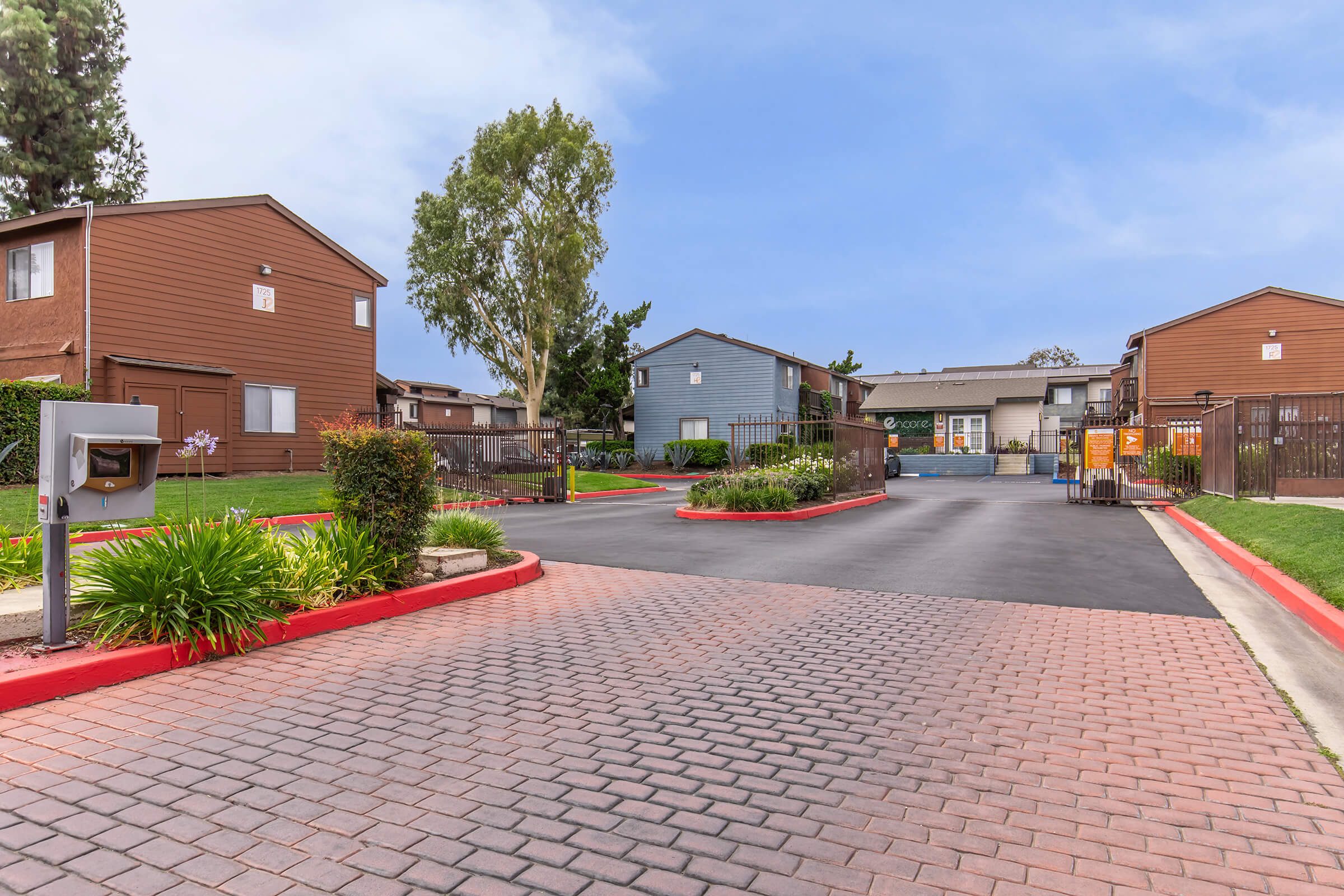 A view of a residential complex featuring two-story brown and gray buildings. The entrance includes a paved driveway with red curbs, greenery along the sides, and a mailbox cluster. In the background, there is a fenced area with additional buildings and amenities. The sky is bright and partly cloudy.