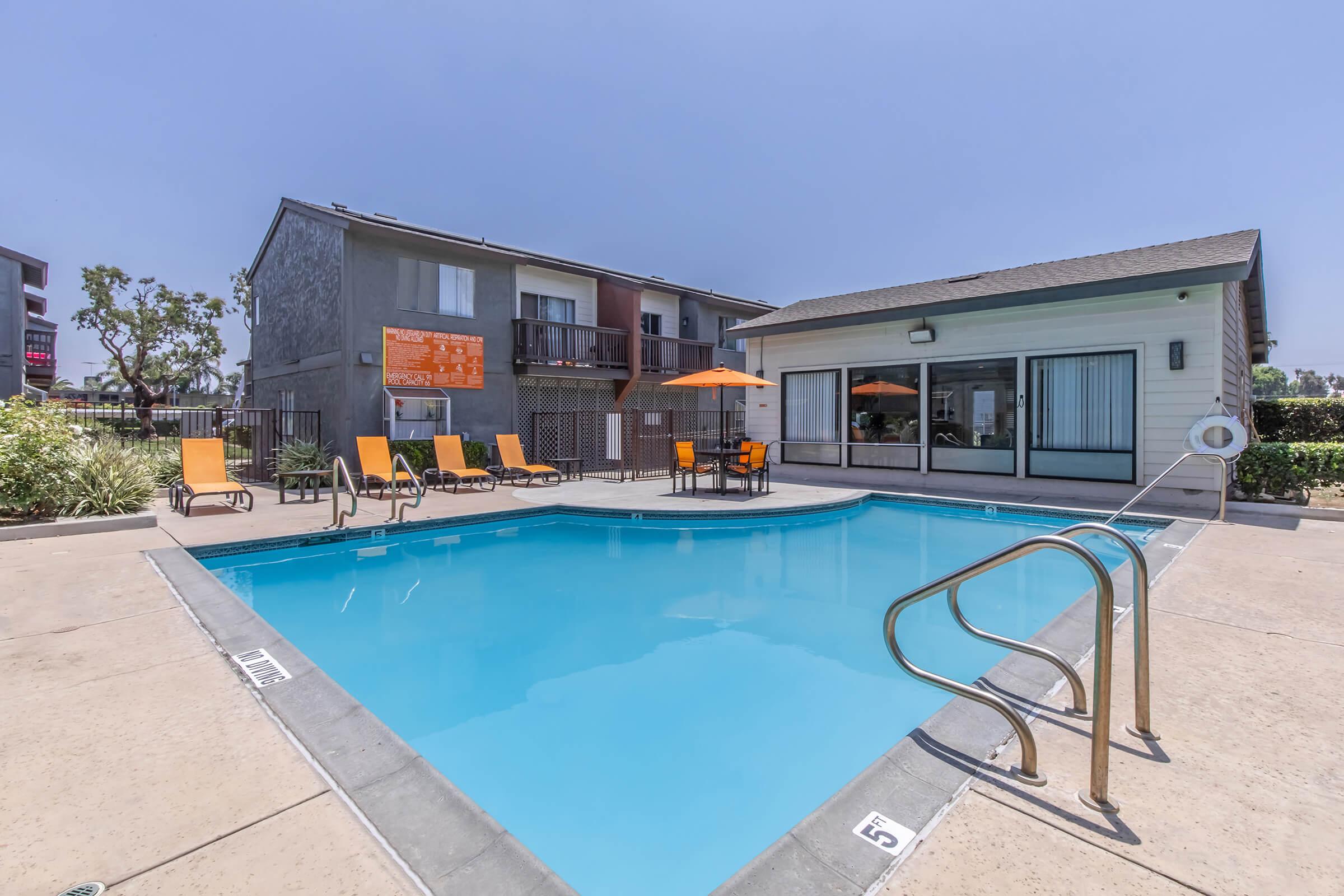 A clear swimming pool surrounded by orange lounge chairs and an umbrella. Nearby is a building with large windows and outdoor seating. The area is sunny, with landscaped greenery in the background.