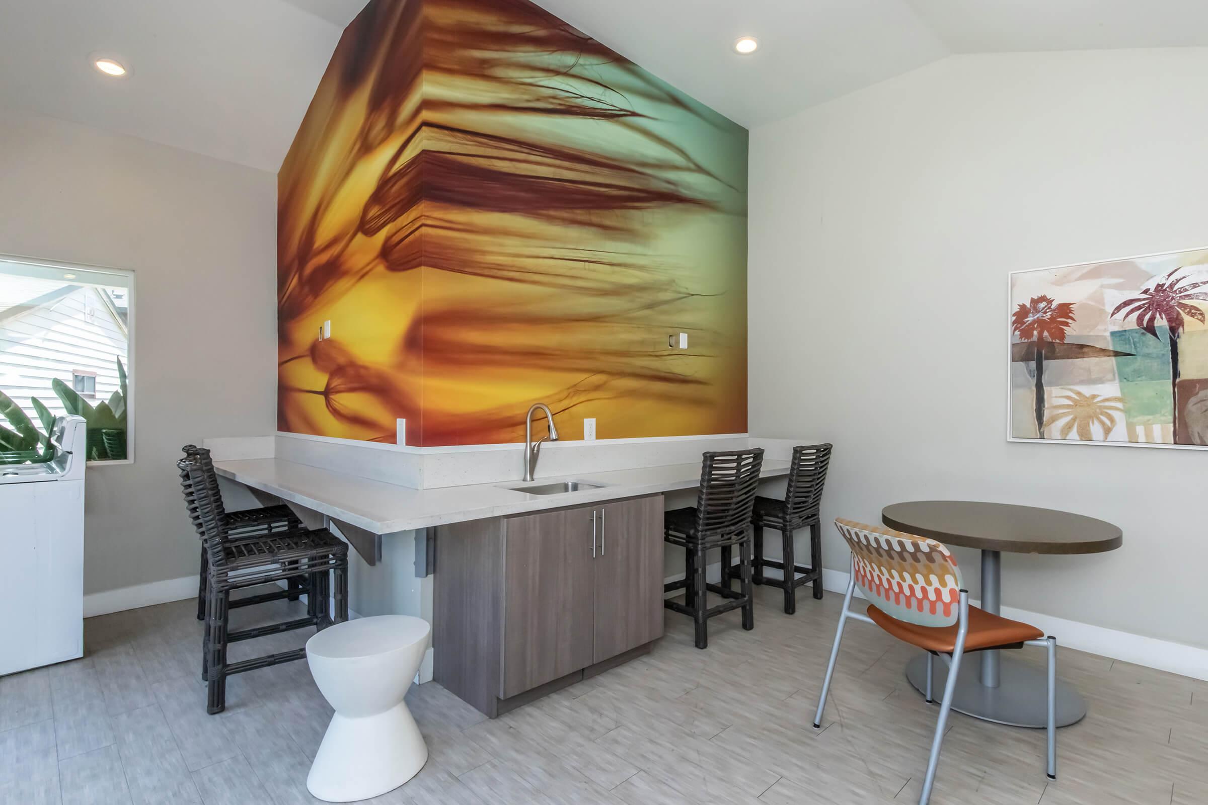 A bright and modern kitchenette featuring a large abstract mural on one wall. The area includes a bar with several black stools, a sleek countertop with a sink, a round table with colorful chairs, and large windows allowing natural light. The decor is minimalist, creating an inviting atmosphere.