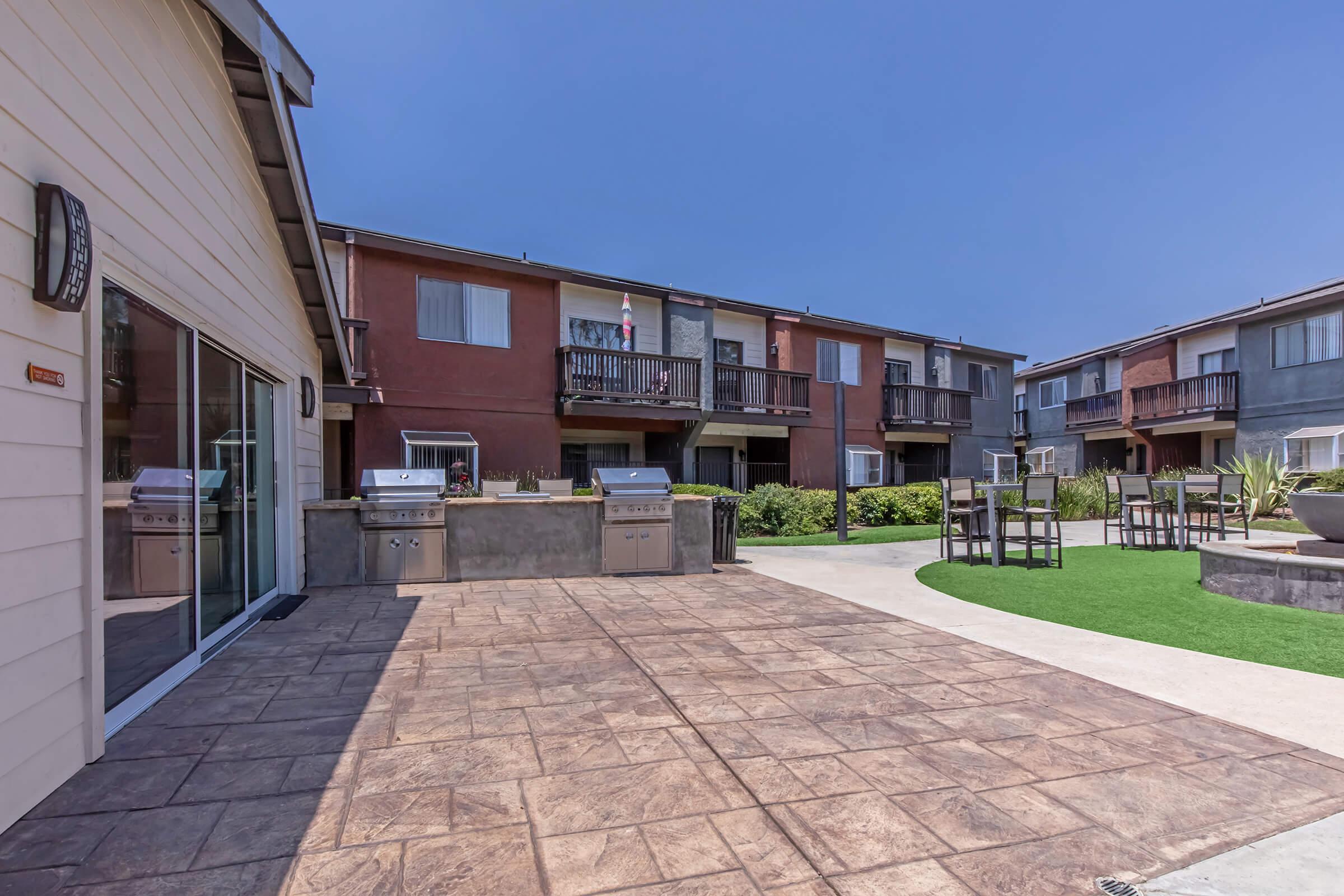 Outdoor area featuring a patio with a textured concrete surface, barbecue grills, and a seating area. In the background, there are two-story apartment buildings with balconies. The space is well-maintained with greenery and a clear blue sky above.
