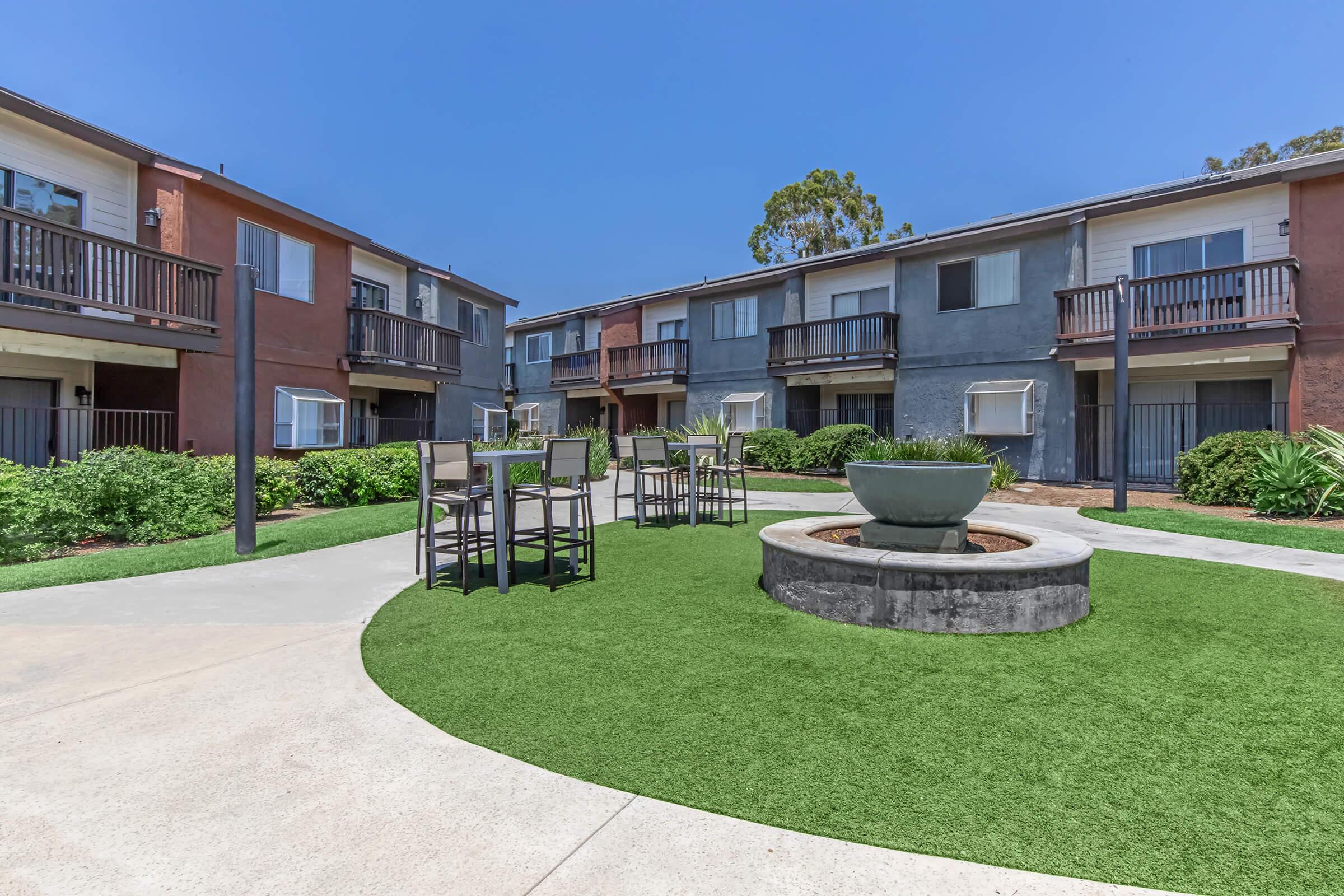 A landscaped courtyard featuring a circular fountain surrounded by green grass. Nearby, there are outdoor tables and chairs. The setting includes two-story apartment buildings with balconies and a clear blue sky in the background.