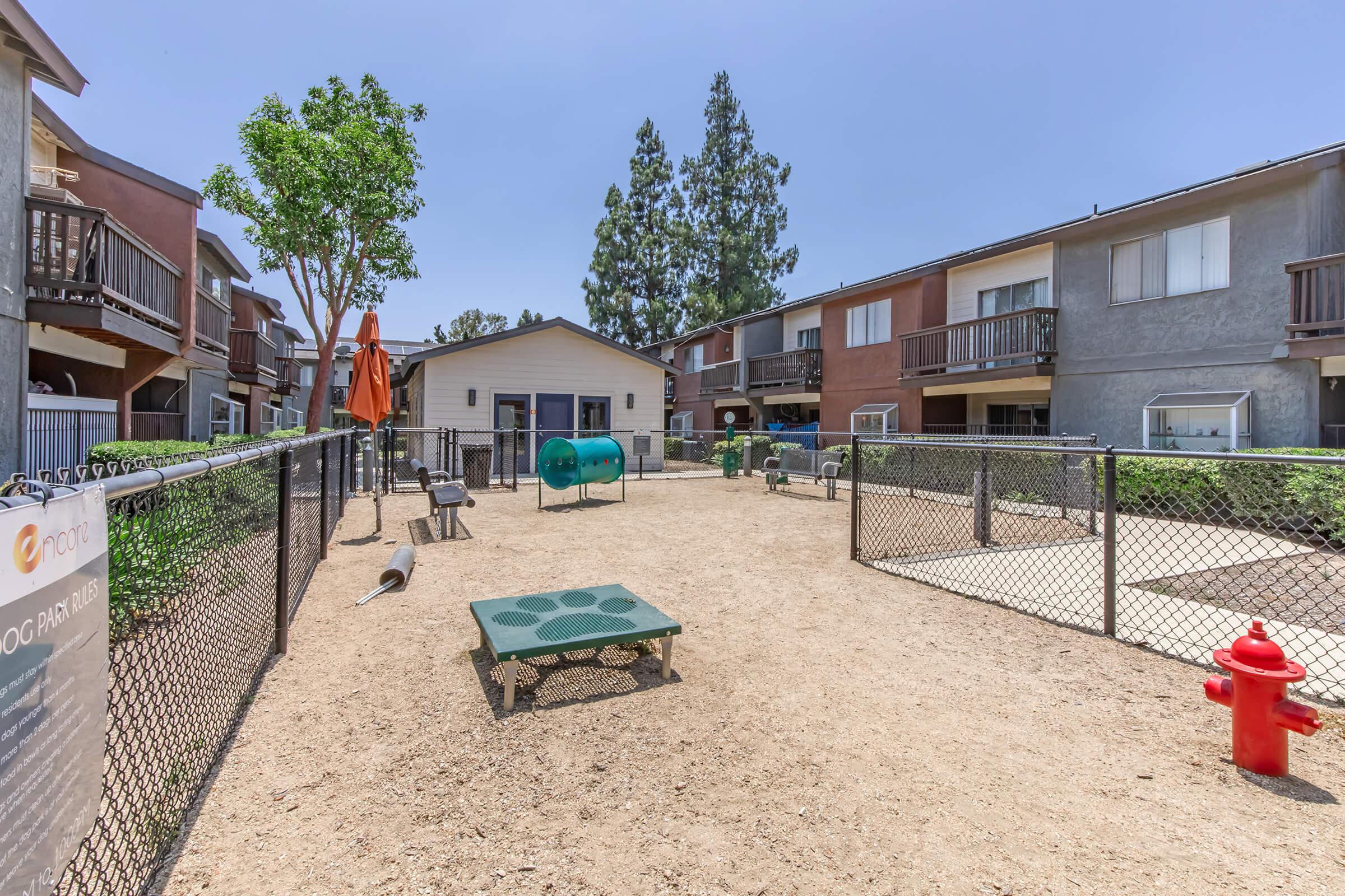 A dog park area featuring a sandy ground enclosed by a chain-link fence. There are picnic tables, a dog agility tunnel, and a red fire hydrant. Surrounding the park are several multi-story residential buildings and trees in a clear blue sky.