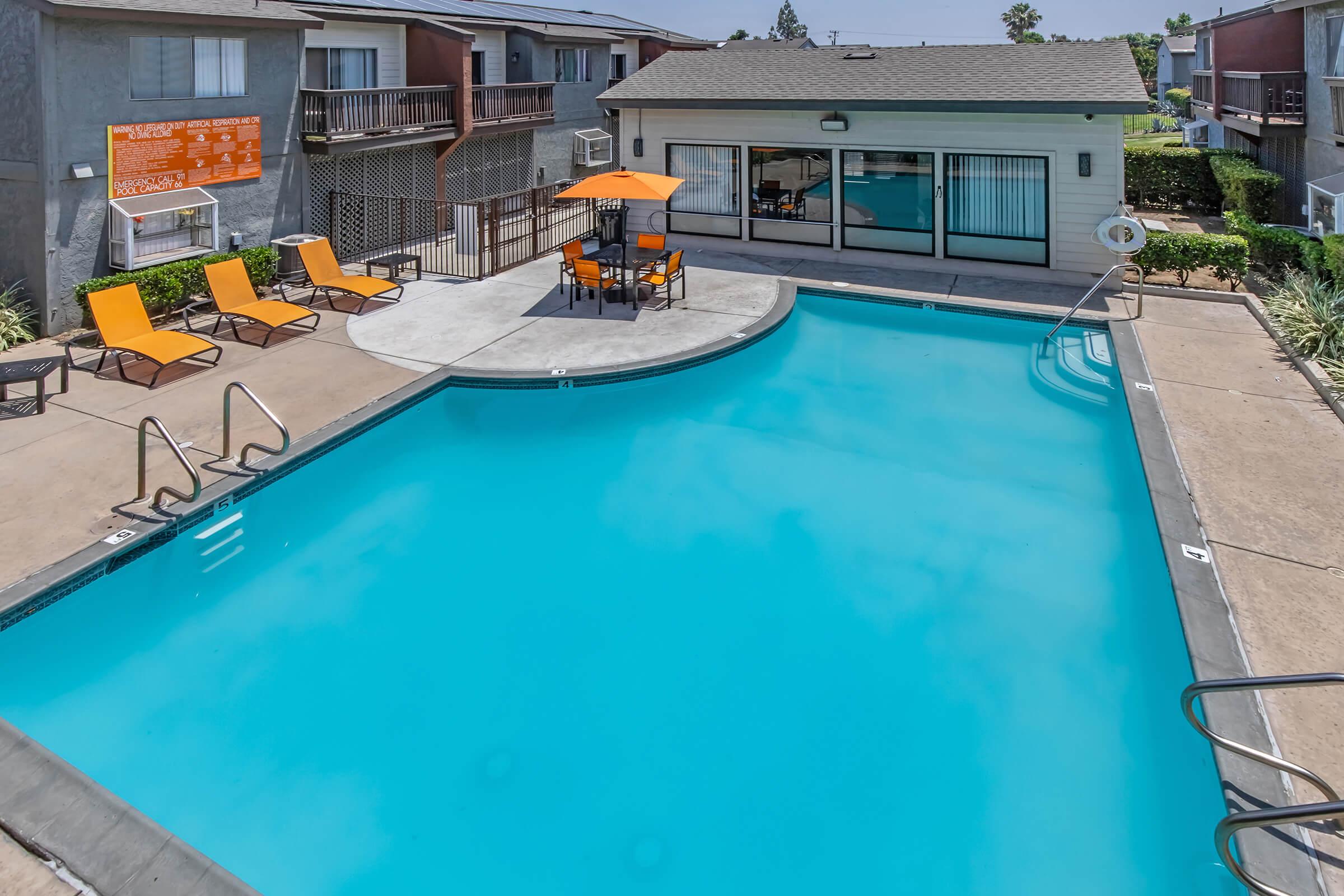 A bright blue swimming pool surrounded by lounge chairs and an orange umbrella. Nearby, there is a small seating area with an orange table and chairs. The background features a multi-story apartment complex with balconies and a clear blue sky above.