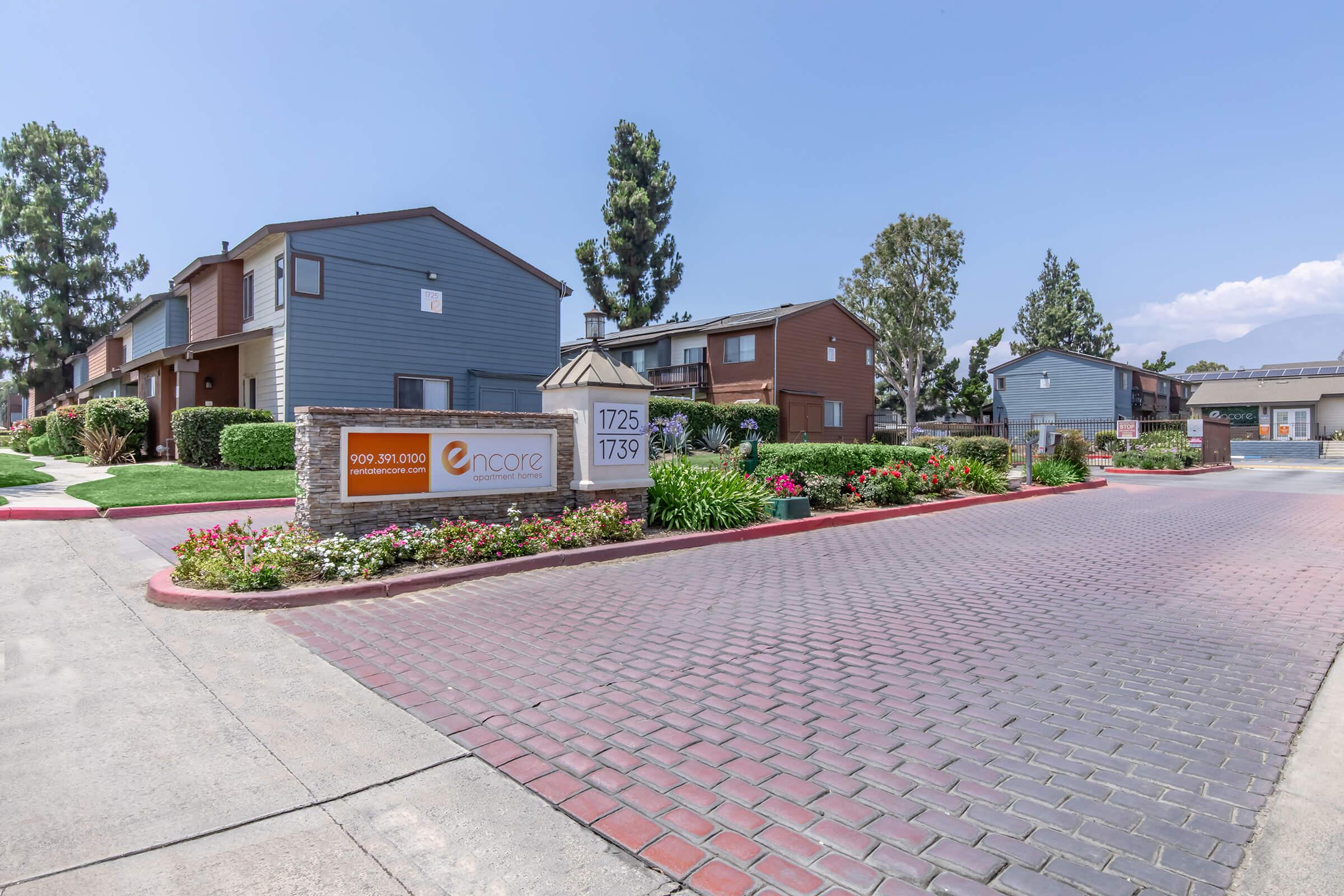 A welcoming entrance to the Encore apartment complex, featuring a stone sign with the name "Encore" and contact information. The path is lined with colorful flower beds, leading to two residential buildings amid well-maintained trees and landscaping under a clear blue sky.
