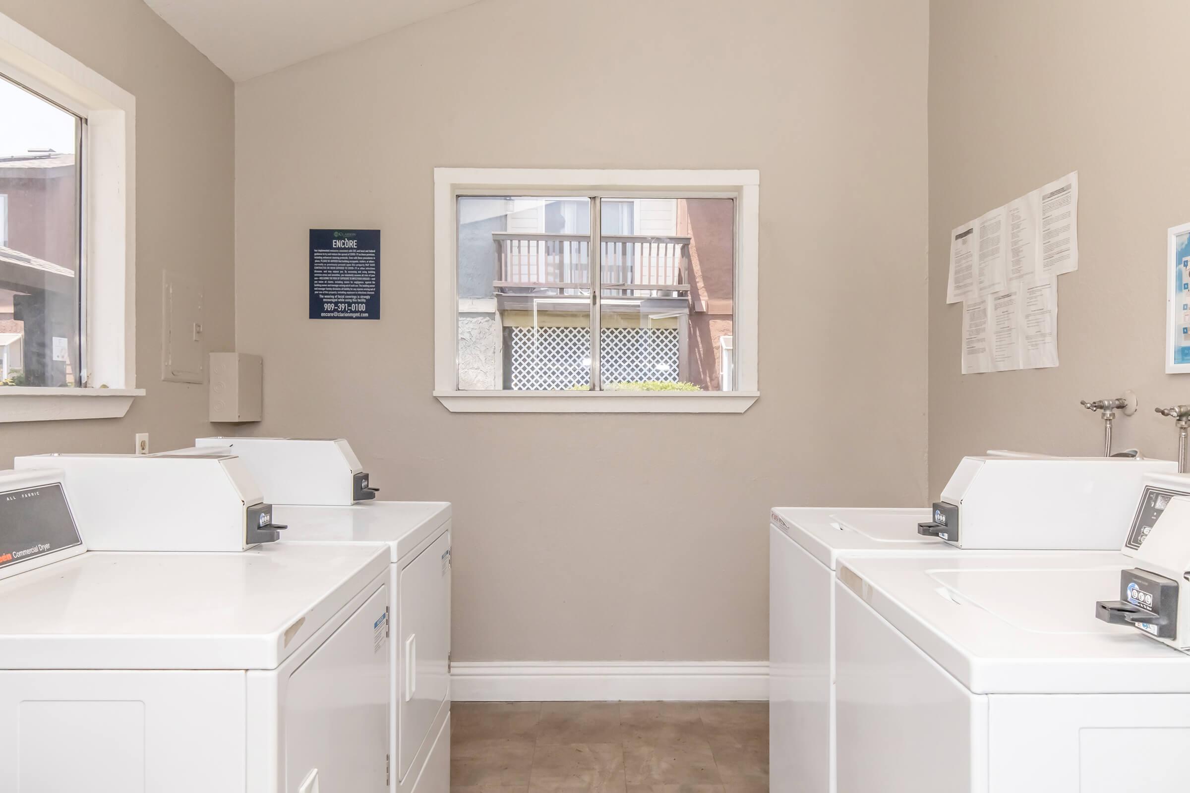 A clean, well-lit laundry room featuring several white washing machines and dryers. There is a large window with a view of a balcony and outdoor space. The walls are painted in a neutral color, and there are informational posters on the walls.
