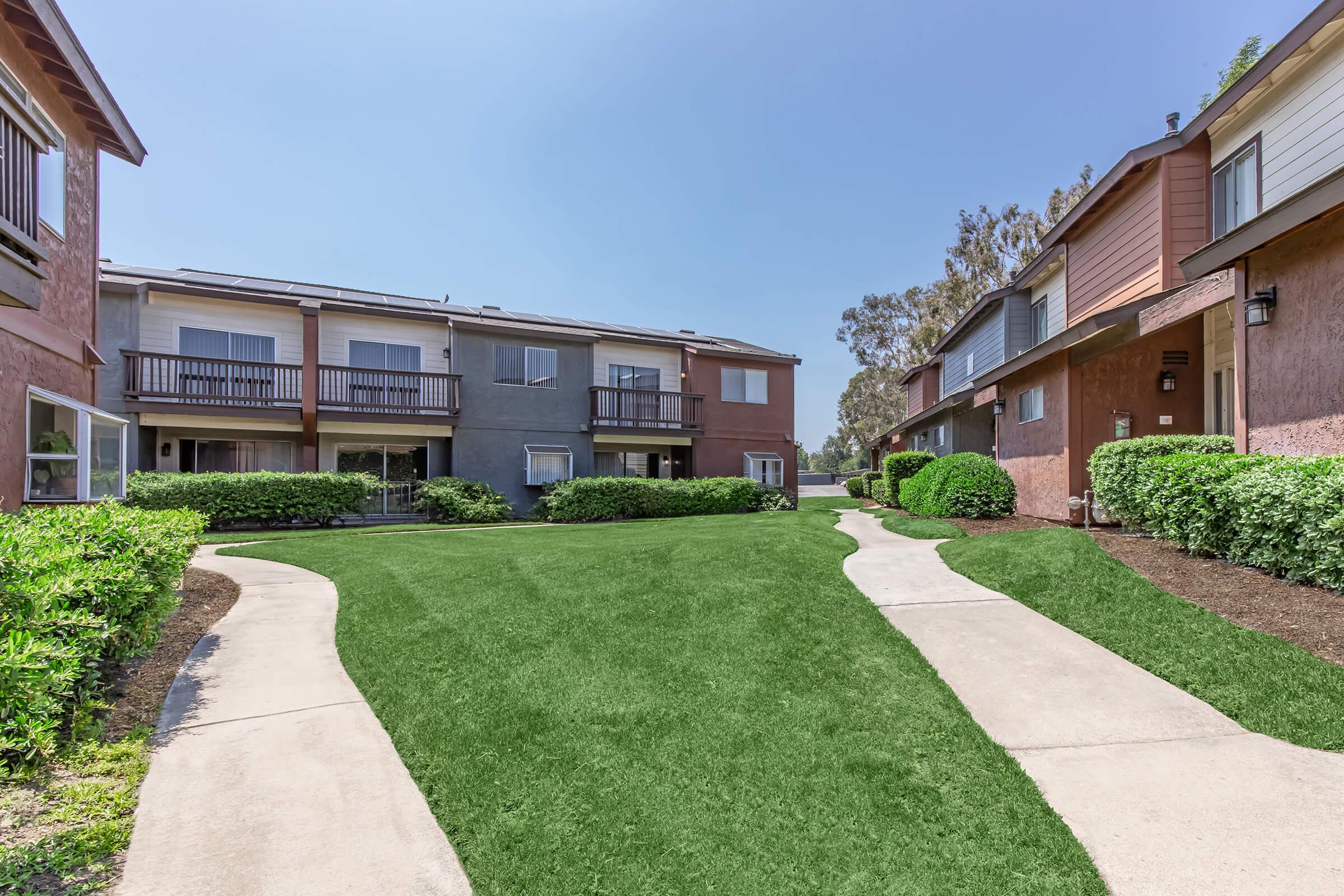 Green lawn area with curved pathways in a residential complex featuring two-story buildings. The buildings have balconies and varied exterior colors, surrounded by neatly trimmed bushes and trees under a clear blue sky.
