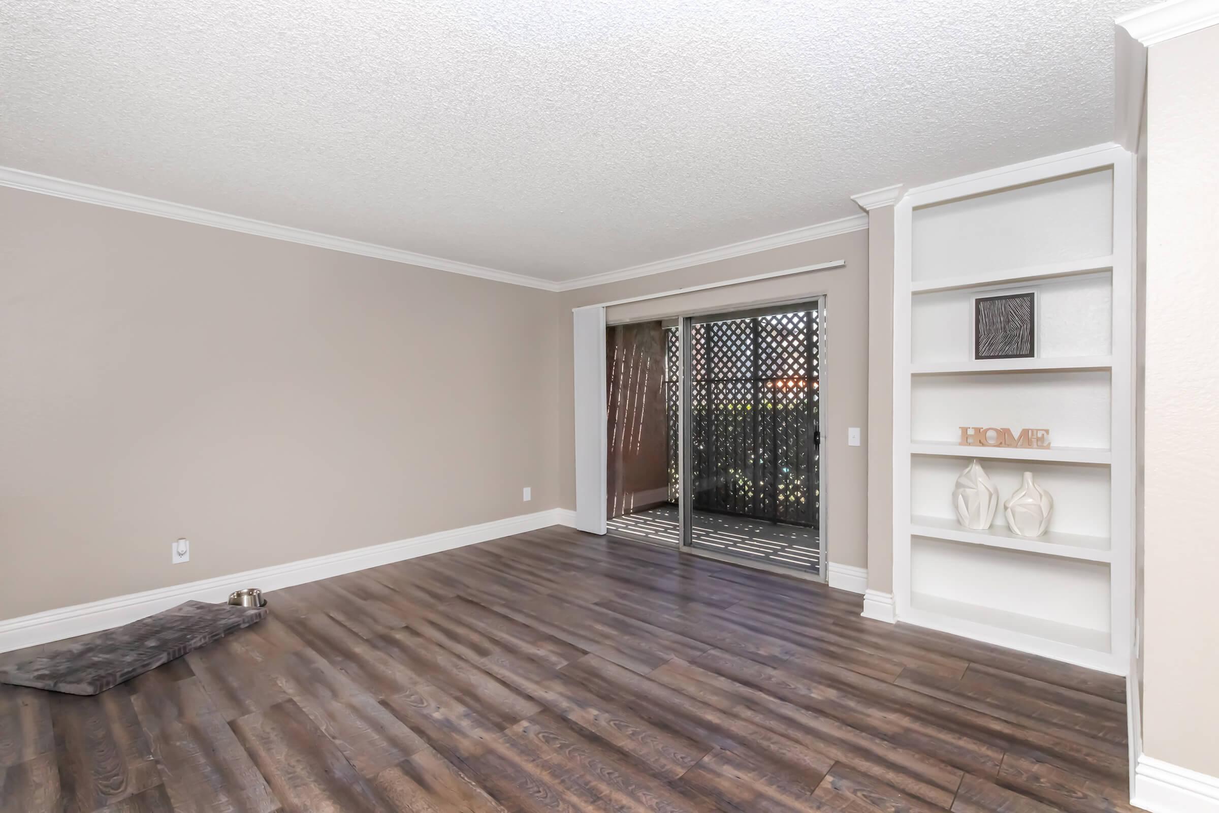 Spacious living room with light-colored walls and dark wooden flooring. A sliding glass door leads to an outdoor area, partially covered by a decorative screen. There is a small pet bed in one corner and a built-in shelf on the adjacent wall displaying decorative objects. Bright and inviting atmosphere.