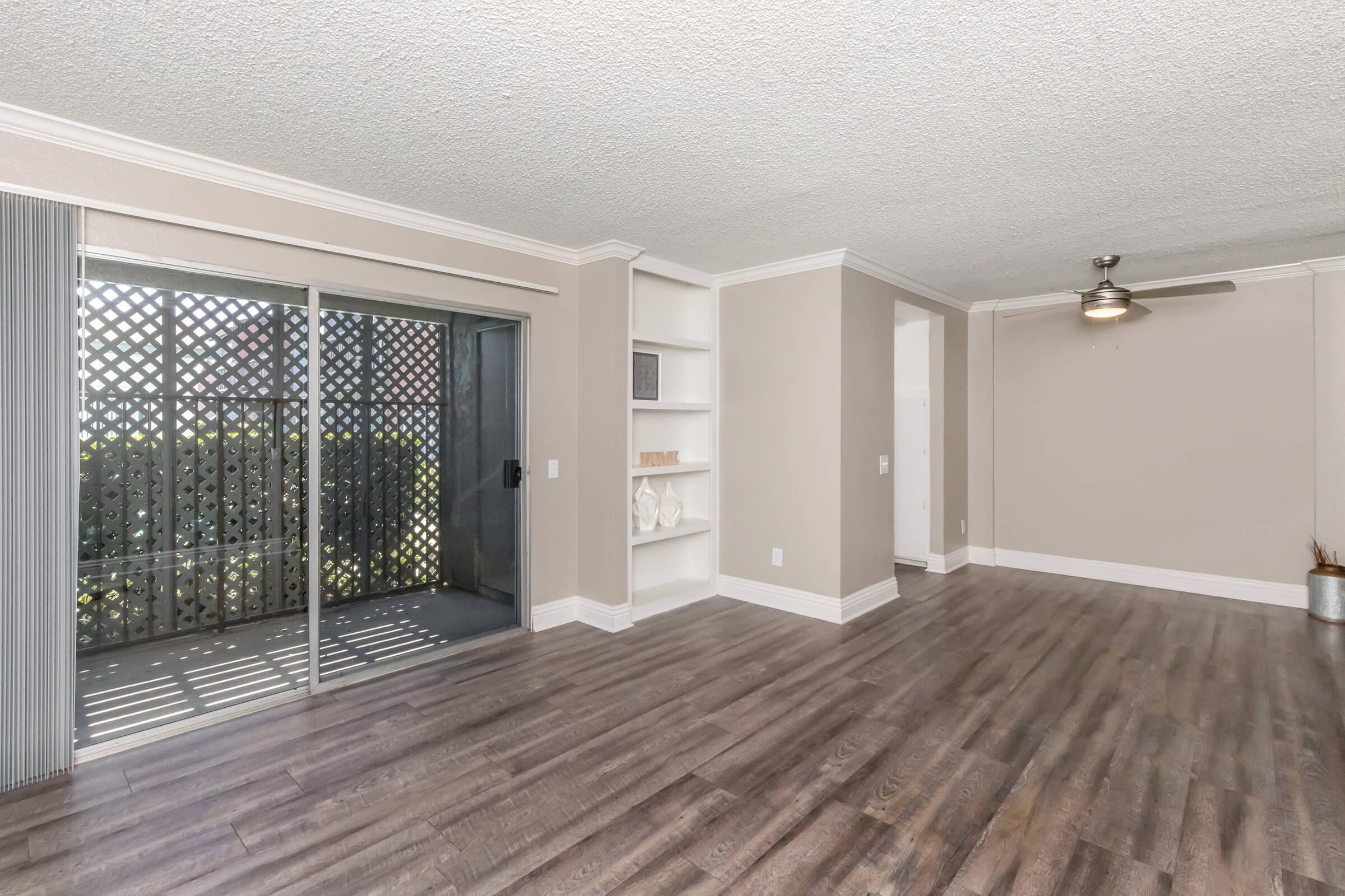 Bright, spacious living area with wooden flooring, featuring large sliding glass doors leading to a balcony. Light neutral-colored walls with a textured ceiling and a ceiling fan. Open shelving built into the wall, providing a modern aesthetic, and ample natural light illuminating the space.