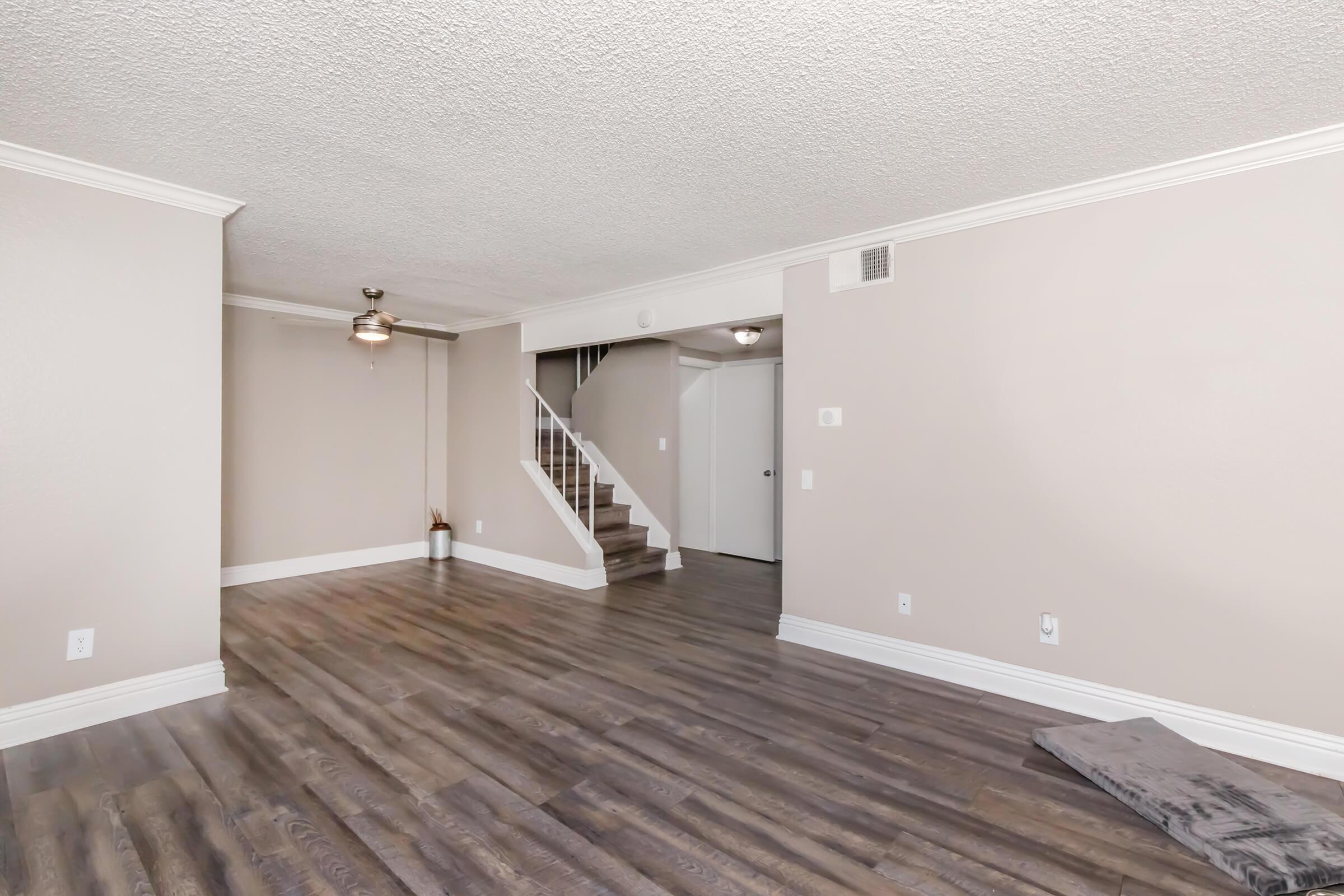 Spacious interior view of a light-colored living room featuring wooden flooring. There is a ceiling fan, a staircase leading up to another level, and walls painted in a neutral shade. A gray mat is placed on the floor near the wall, and a closed door is visible in the background.