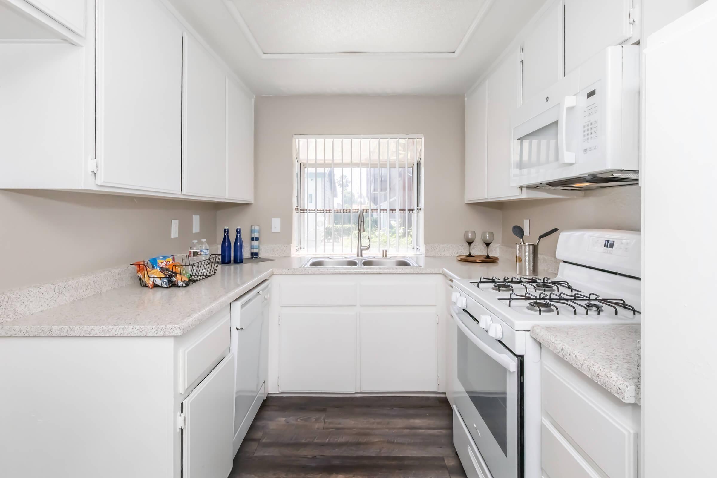 A bright kitchen featuring white cabinetry, a countertop, stainless steel appliances, and a window providing natural light. The space includes a sink, a stove, and decorative items like wine glasses and blue bottles on the countertop, creating a clean and inviting atmosphere.