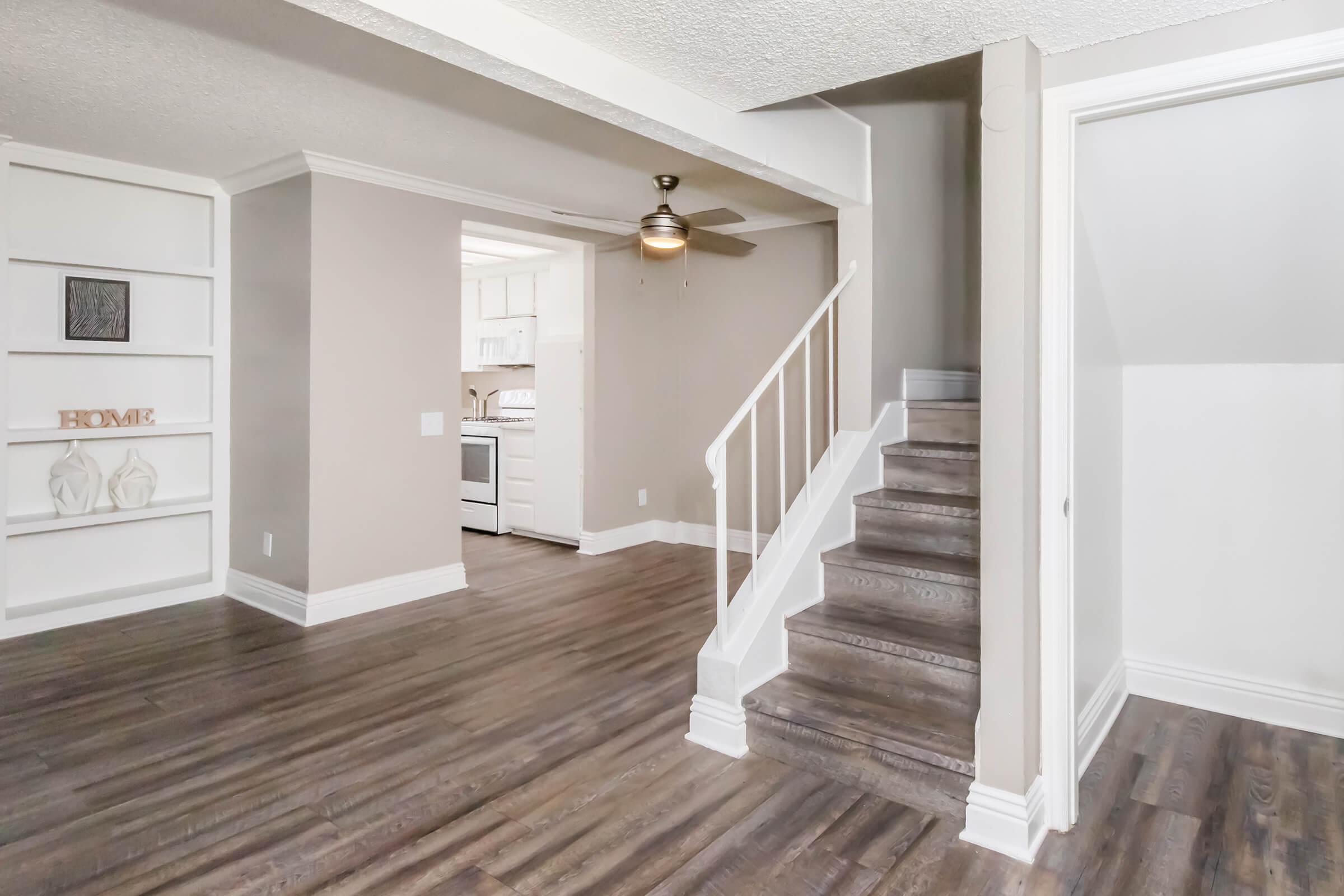 A spacious interior view featuring a living area with wood flooring, a ceiling fan, and a nearby staircase leading to an upper level. The walls are painted in neutral tones, and there are built-in shelves on one side. A doorway opens to a kitchen area visible in the background.
