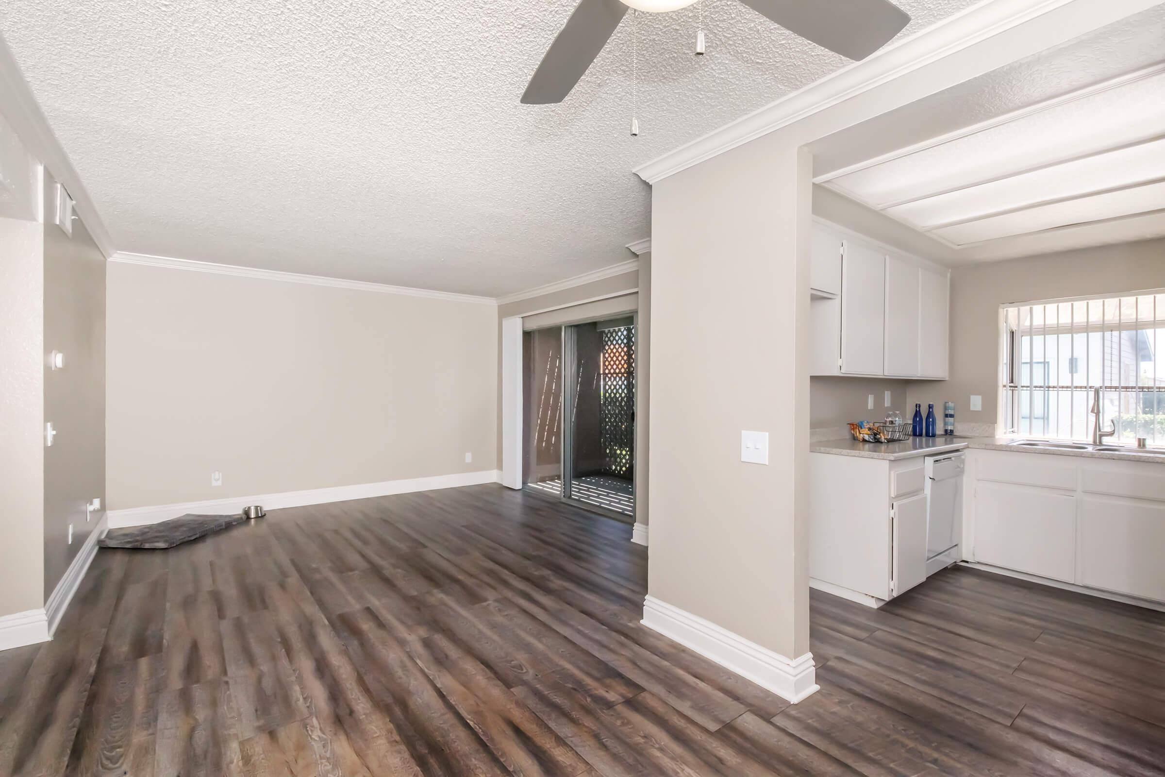 Empty living space featuring light beige walls, a ceiling fan, and hardwood-style flooring. To the left, there's a small area suitable for a pet bed, and on the right, an open kitchen with white cabinetry and a window overlooking an outdoor area.