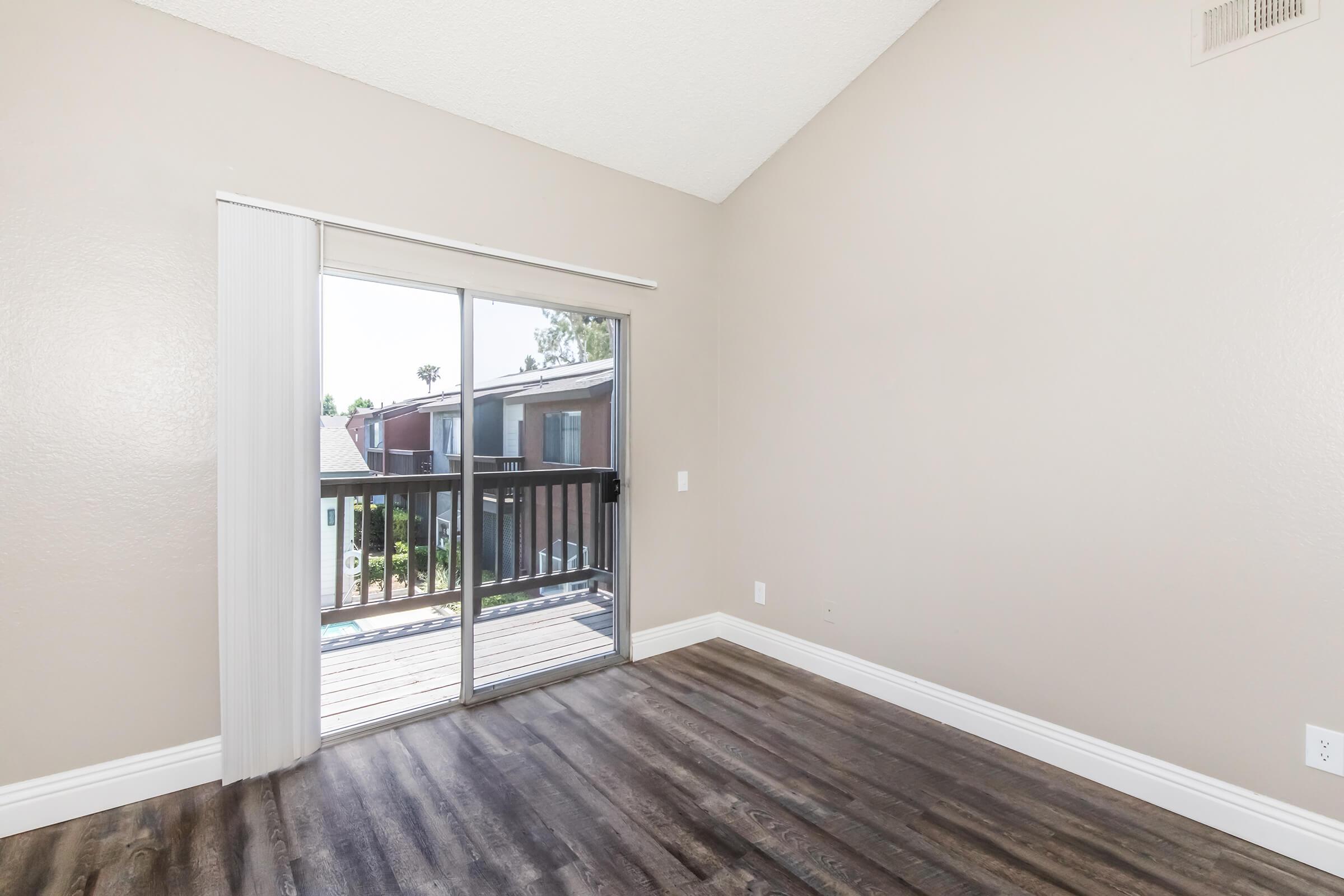 Empty room with light beige walls and dark wooden flooring. A sliding glass door opens to a small balcony with a view of neighboring buildings. The ceiling is vaulted, creating an airy feel. Natural light streams in through the door, illuminating the space.