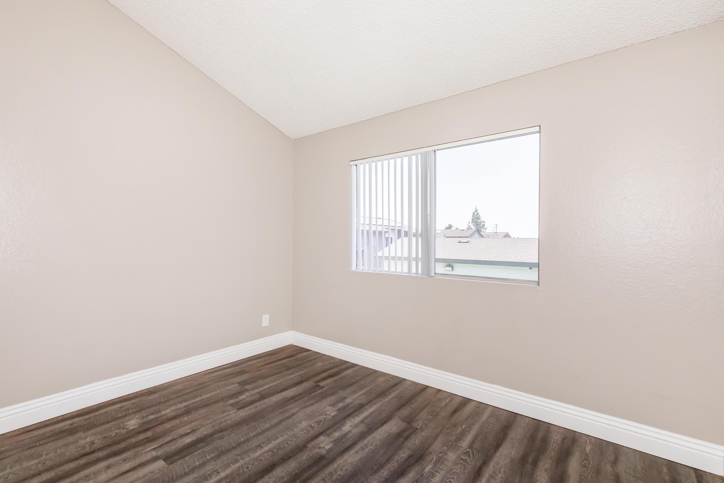 A light, empty room with a sloped ceiling and beige walls. The floor features dark wooden laminate. There is a window with vertical blinds, allowing natural light to enter, and it overlooks a residential area outside.