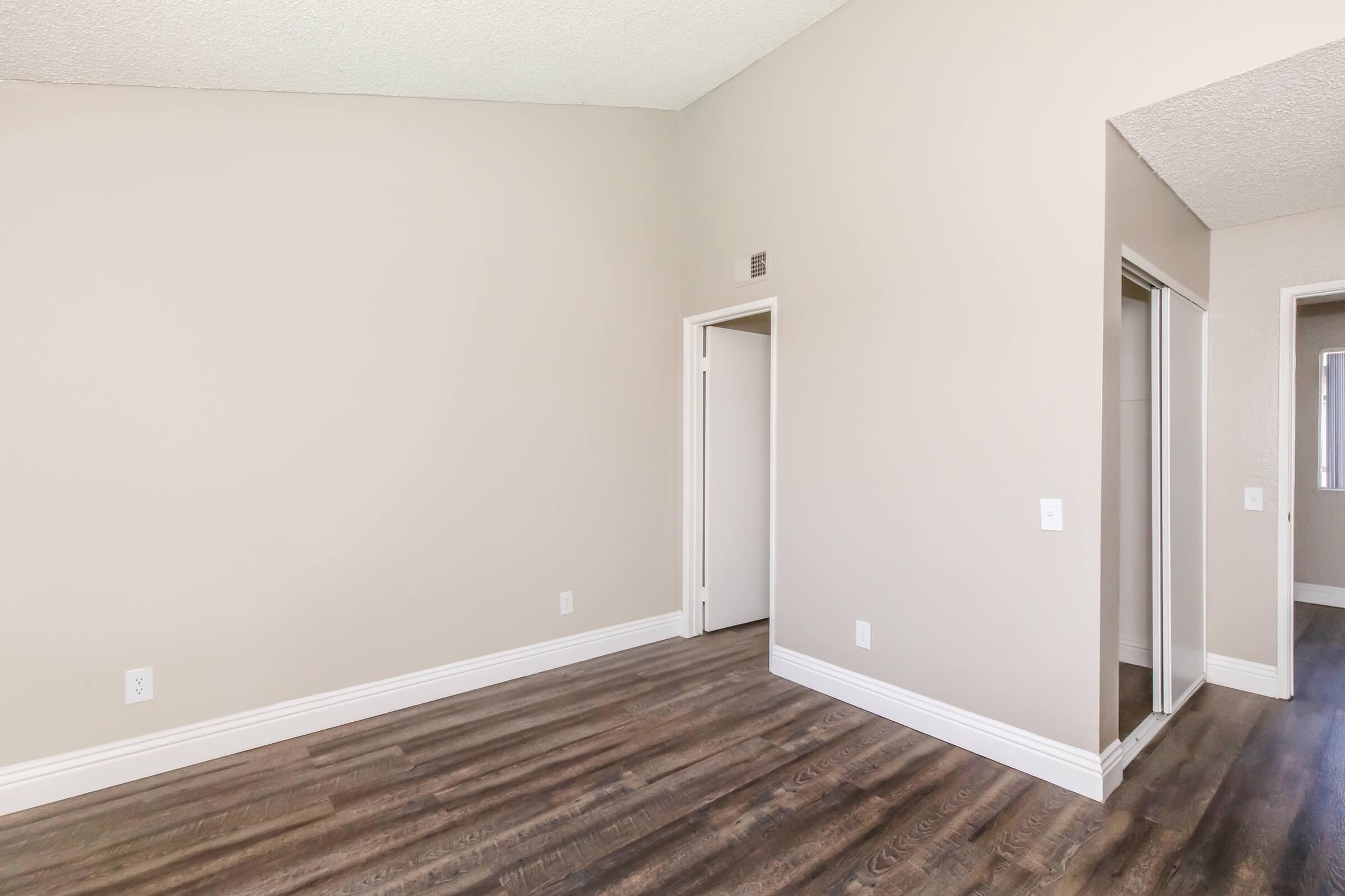 Interior view of a spacious room with light-colored walls and a textured ceiling. The floor is covered in dark wood laminate. There is a doorway leading to another room and a mirrored closet on one side. The overall ambiance is bright and neutral, suitable for various decor styles.