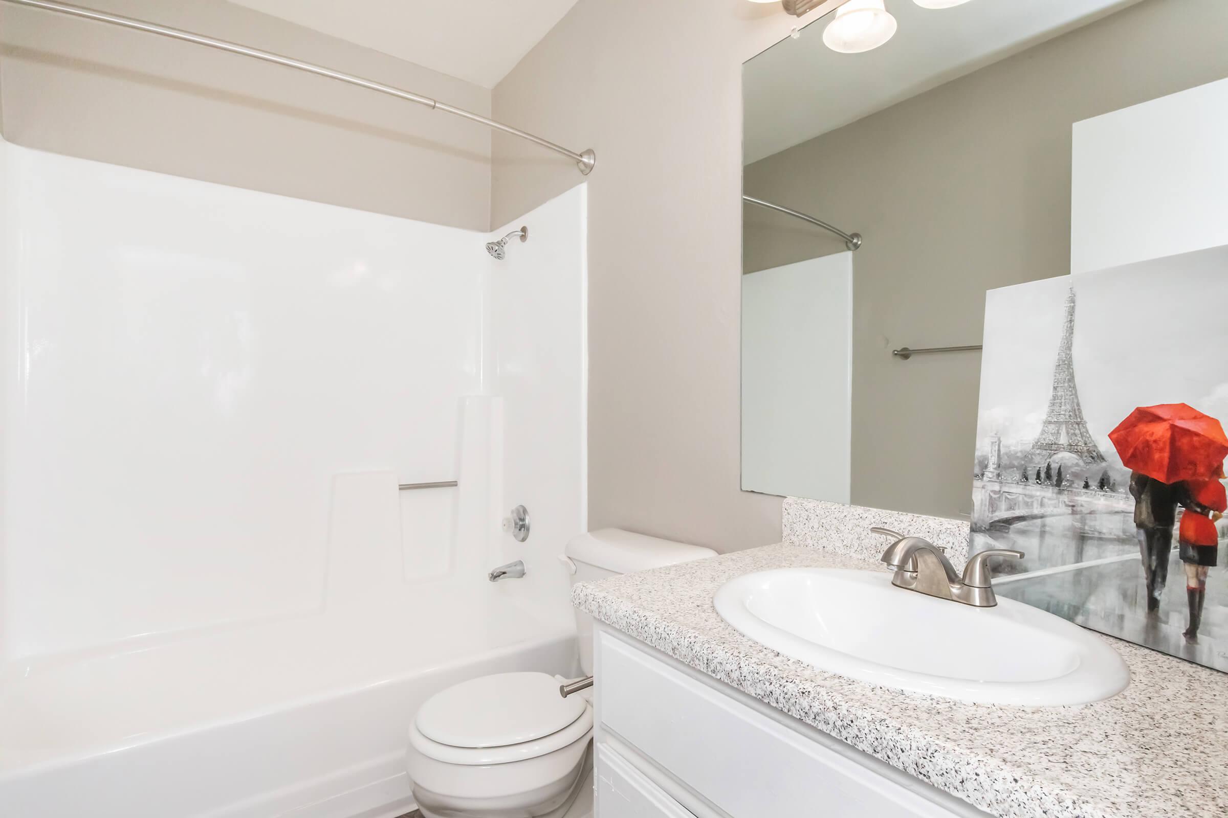 A modern bathroom featuring a white bathtub with a shower, a light granite countertop with a sink, and a mirror. The wall is painted in a neutral color, and there is a decorative image of the Eiffel Tower with a couple holding red umbrellas leaning against the countertop.