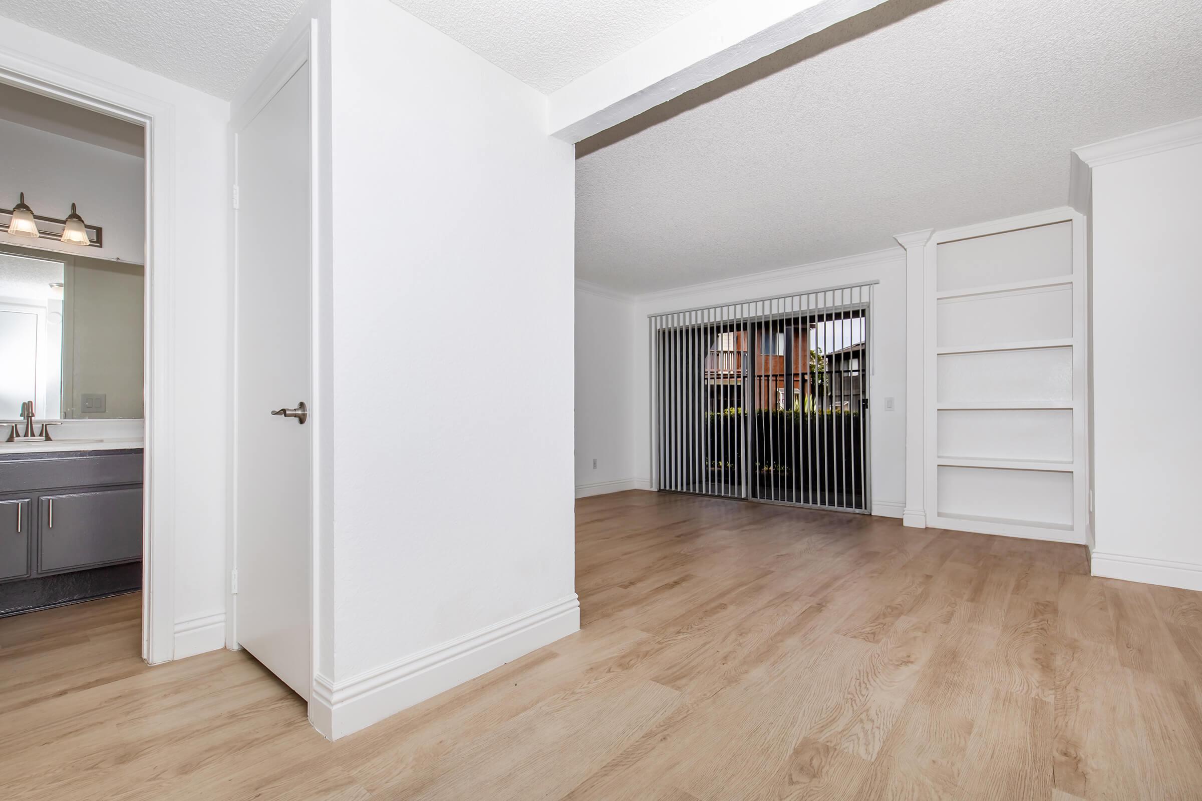 A bright, modern living space featuring light-colored walls and wooden flooring. On the left, there is a doorway leading to a bathroom with dual sinks. The right side showcases a wall with built-in shelves and a large sliding door with vertical blinds that opens to an outdoor area.