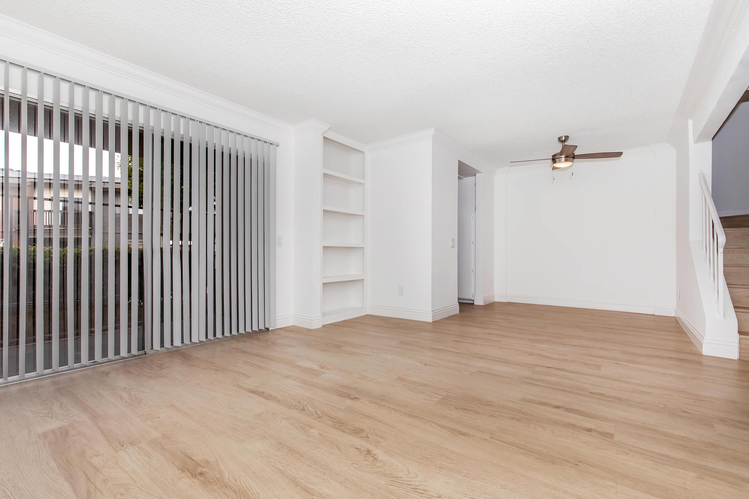 A spacious, empty living room with light hardwood flooring. There are vertical blinds covering a large sliding glass door, and a ceiling fan is mounted above. White walls and built-in shelves add to the minimalistic design. No furniture is present, giving it a clean and open feel.