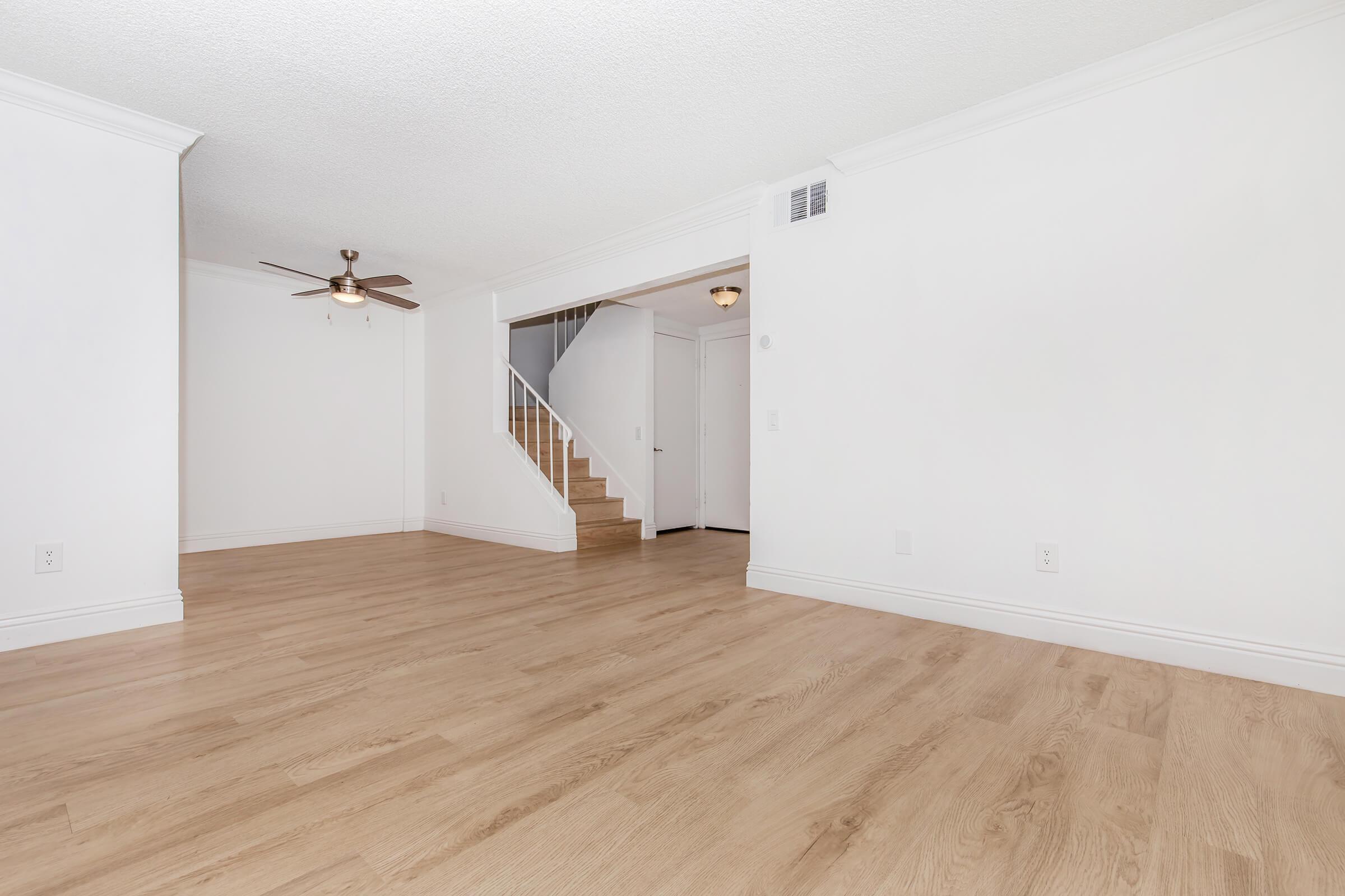 Spacious, empty living room featuring light wood flooring, a ceiling fan, and a staircase leading to an upper level. The walls are painted white, providing a bright and open atmosphere. A doorway to the right leads to another room or hallway.