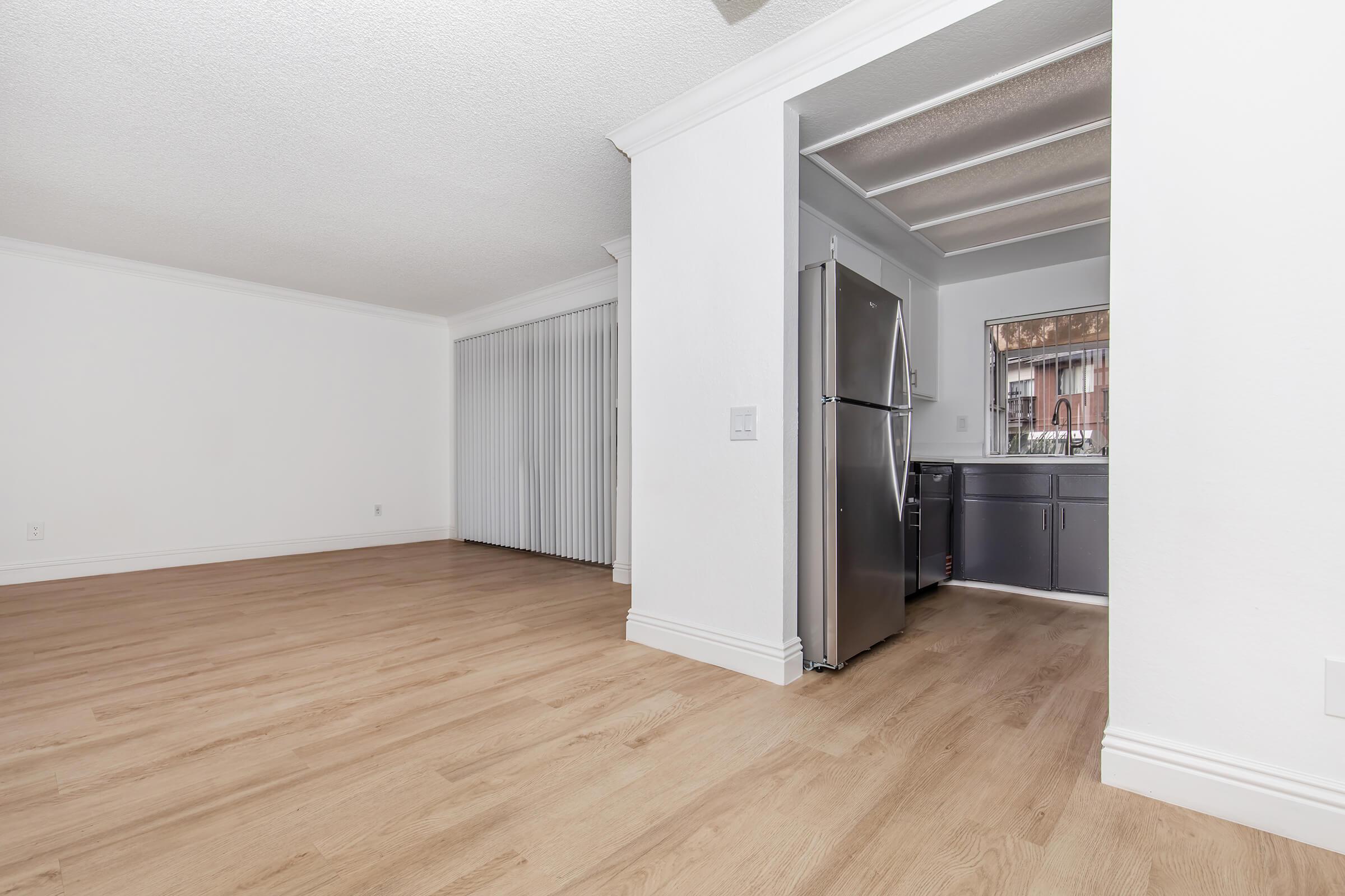 A spacious and bright room with light-colored walls and wooden flooring. To the right, there is a kitchen area featuring a stainless steel refrigerator and dark cabinets. Large windows allow natural light, and there are vertical blinds on the windows in the background. The space appears clean and ready for furnishing.