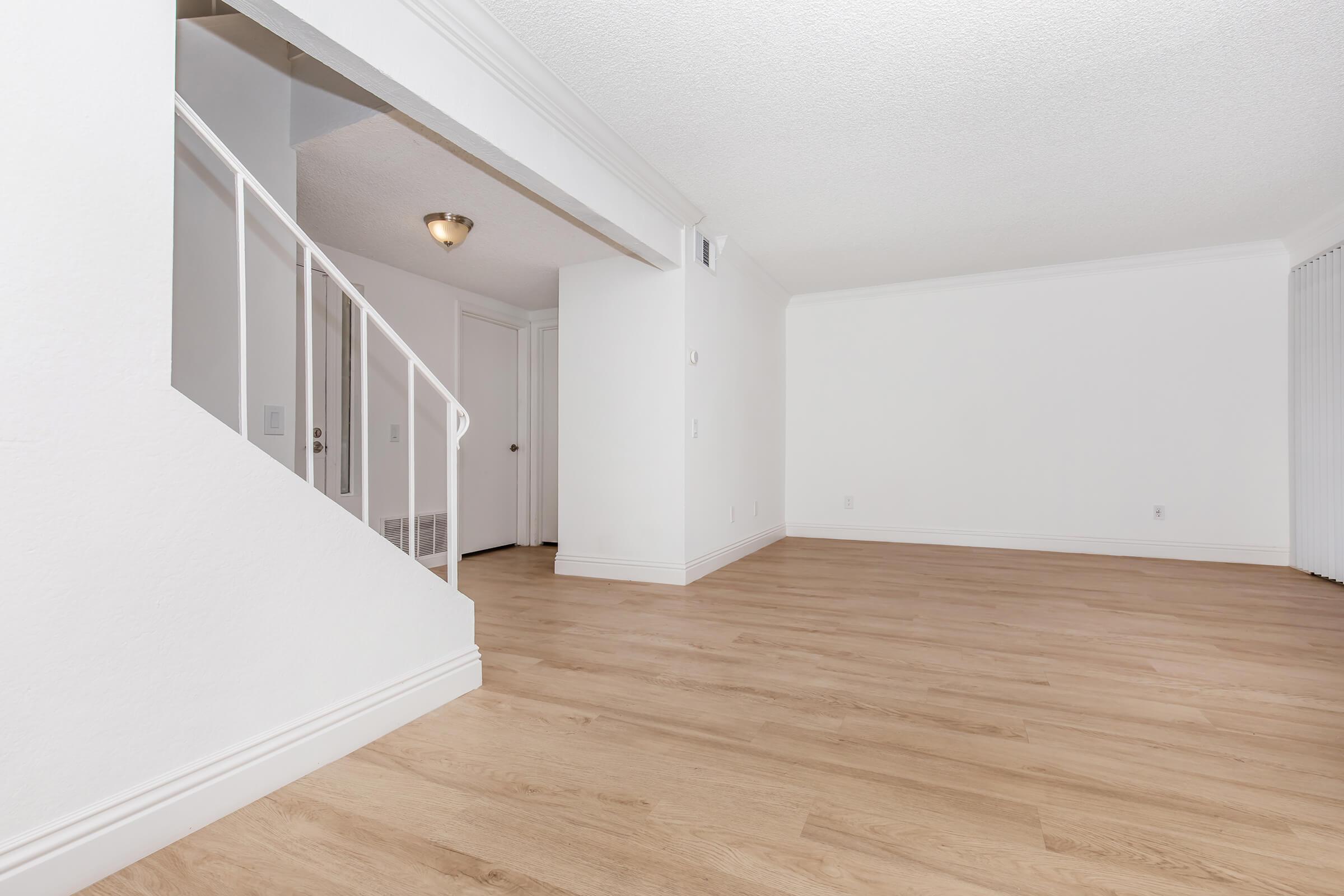 Bright and empty living space featuring light wood flooring, white walls, and a staircase. Natural light enters from the left, highlighting the clean and simple design of the room, which is ready for furniture placement.
