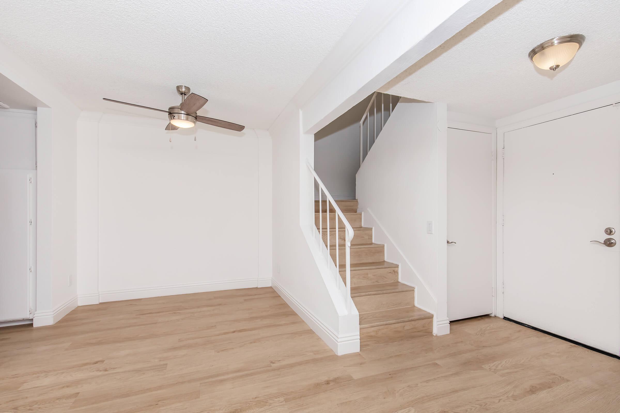 A bright and spacious interior view of a modern home. The room features light wooden flooring, a ceiling fan, and white walls. A staircase leads to the upper level, and a door is visible at the entrance, enhancing the open and airy feel of the space. Natural light fills the area, creating a welcoming atmosphere.