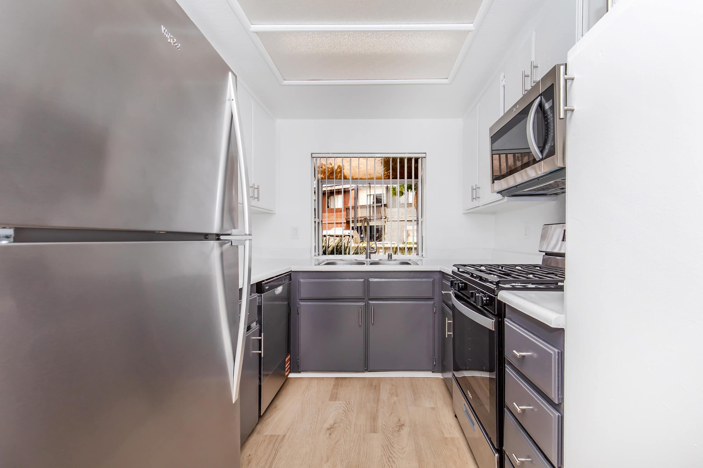 A modern kitchen featuring stainless steel appliances, including a refrigerator, oven, and microwave. The kitchen has gray cabinets, light wood flooring, and a window with vertical blinds allowing natural light. The layout is compact, emphasizing functionality and contemporary design elements.