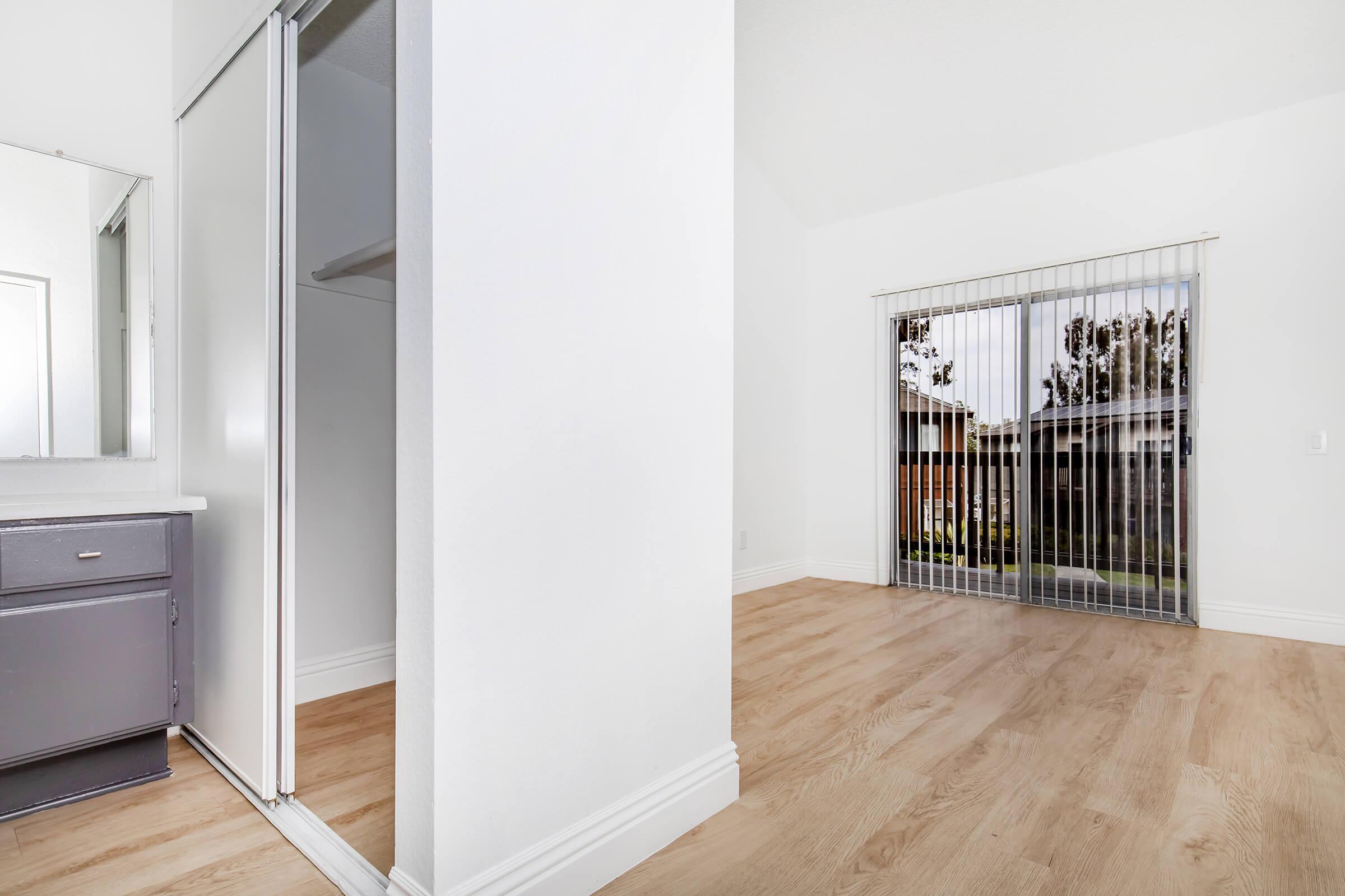 Interior view of a bright room featuring light wood flooring, a mirrored closet on one side, and large sliding glass doors leading to an outdoor space. The walls are painted white, and there is a spacious, airy feel with natural light coming through the windows.