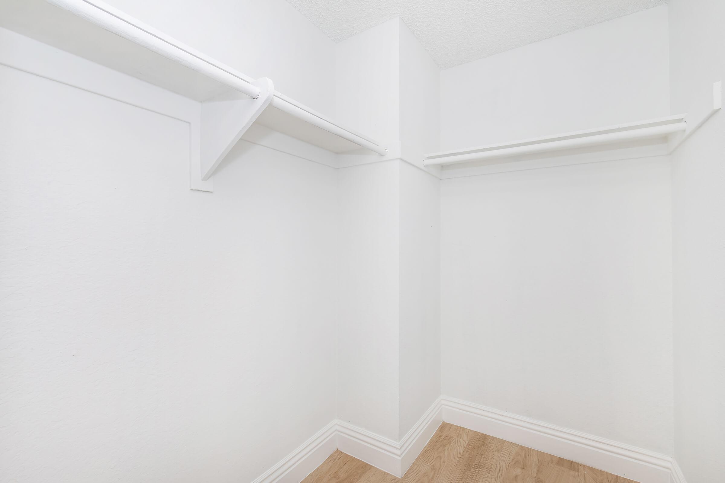 Empty white closet with two hanging shelves and light brown flooring, featuring clean walls and corners. The space is well-lit and free of any items, creating a minimalist storage area.