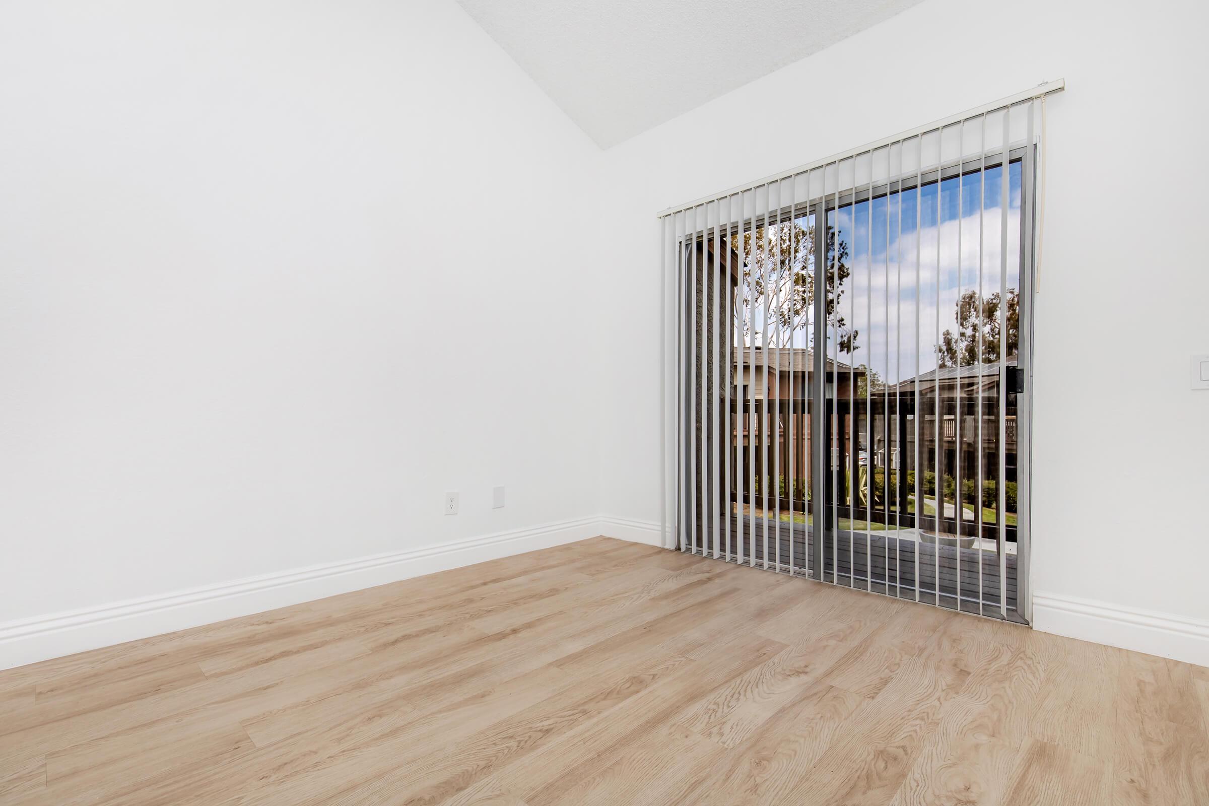 Empty room with light wooden flooring, white walls, and a large window covered with vertical blinds. The window shows a view of trees and a cloudy sky outside, creating a bright and airy atmosphere. The ceiling is high, enhancing the spacious feel of the room.