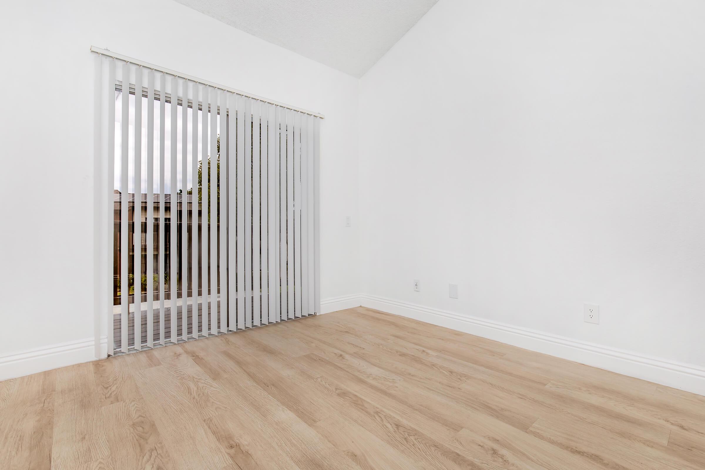A bright, empty room with white walls and wooden flooring. Vertical blinds cover a glass door leading to an outdoor area, allowing natural light to enter. The ceiling is high with a slight slope, creating a spacious feel. The room is uncluttered and ready for furnishing.