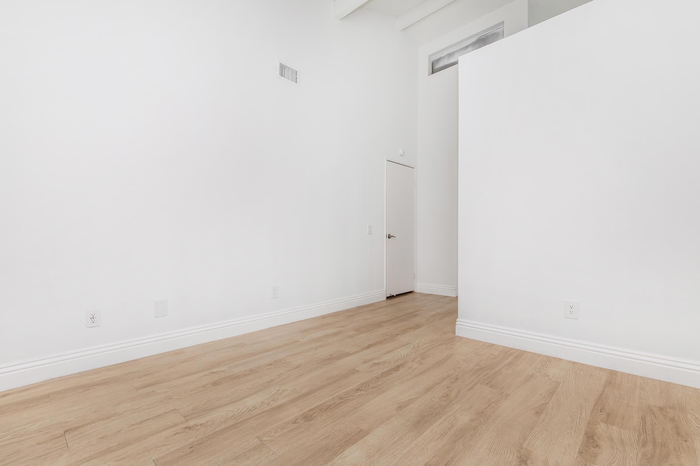 A spacious, empty room with white walls and a light wooden floor. There is a door on the left side and a partial wall on the right, creating a sense of openness and minimalism. The room is well-lit with natural light coming from a window above the door.