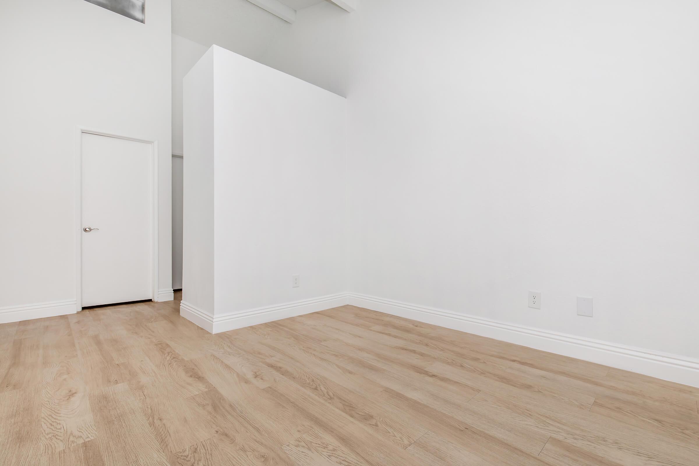 A spacious, empty room with light wood flooring, white walls, and high ceilings. There's a door on one side and a partial wall creating a division within the space. The overall ambiance is bright and minimalistic, suitable for various uses like a living area or office.