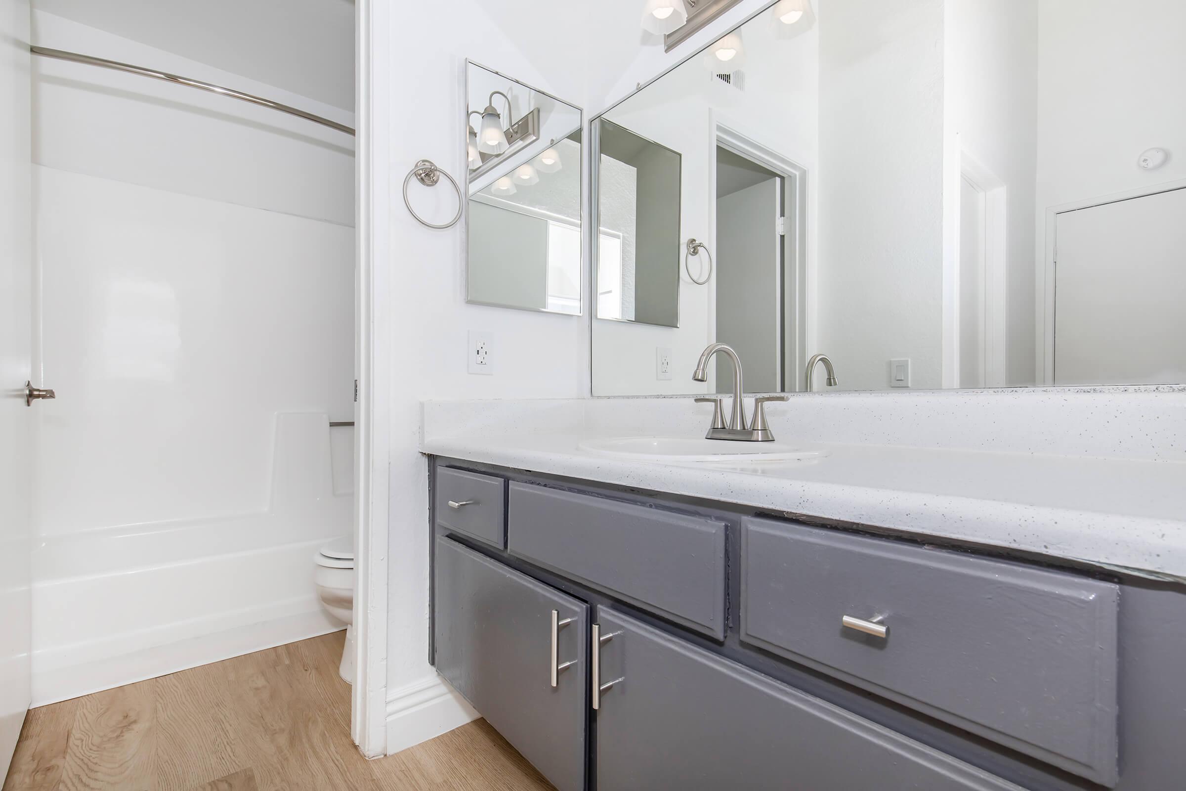 A modern bathroom featuring a clean design with a gray vanity, a white sink, and a large mirror above. The shower area is minimalist with white walls and a simple curved shower rod. Light wood flooring complements the space, contributing to a fresh and airy atmosphere.