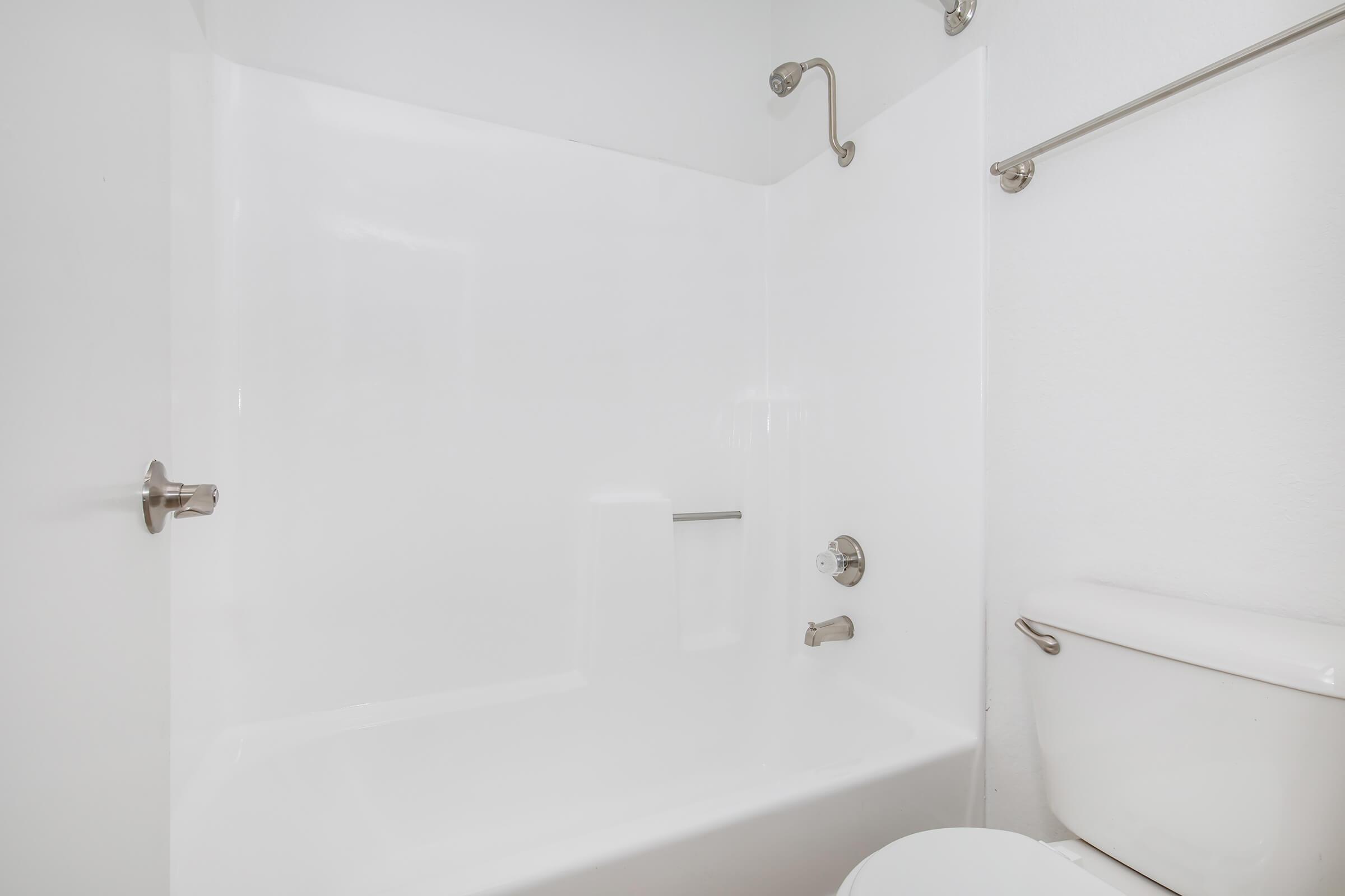 A clean and minimalistic bathroom featuring a white bathtub with a showerhead, a chrome faucet, and a white toilet. The walls and bathtub are also white, creating a bright and open atmosphere. The space is uncluttered, emphasizing a modern design aesthetic.