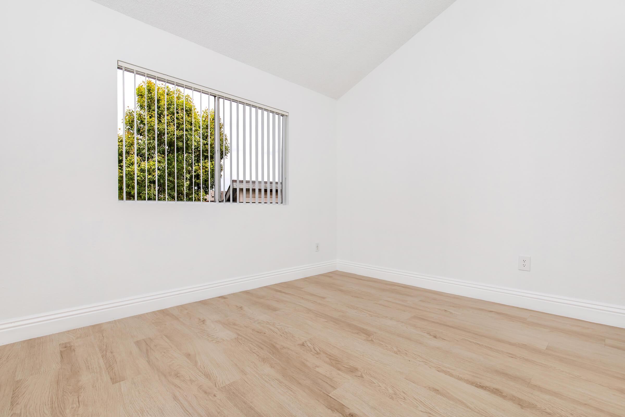 An empty room with light-colored wooden flooring and white walls. A window with vertical blinds allows natural light, revealing a green tree outside. The ceiling is angled, giving a spacious feel to the room, which has no furniture or decorations.