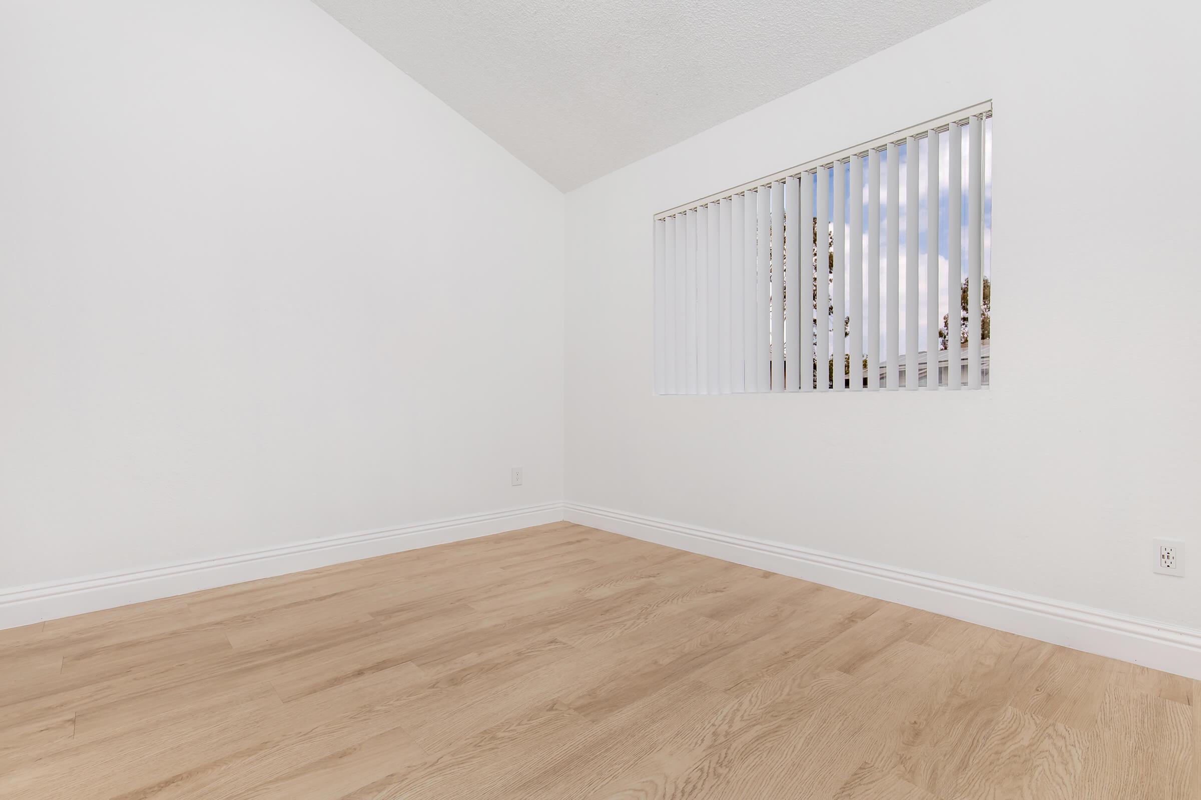 A bright, empty room with a high ceiling and light wood flooring. A window with horizontal blinds allows natural light to enter, illuminating the white walls. The space appears clean and minimalistic, ideal for various interior design possibilities.