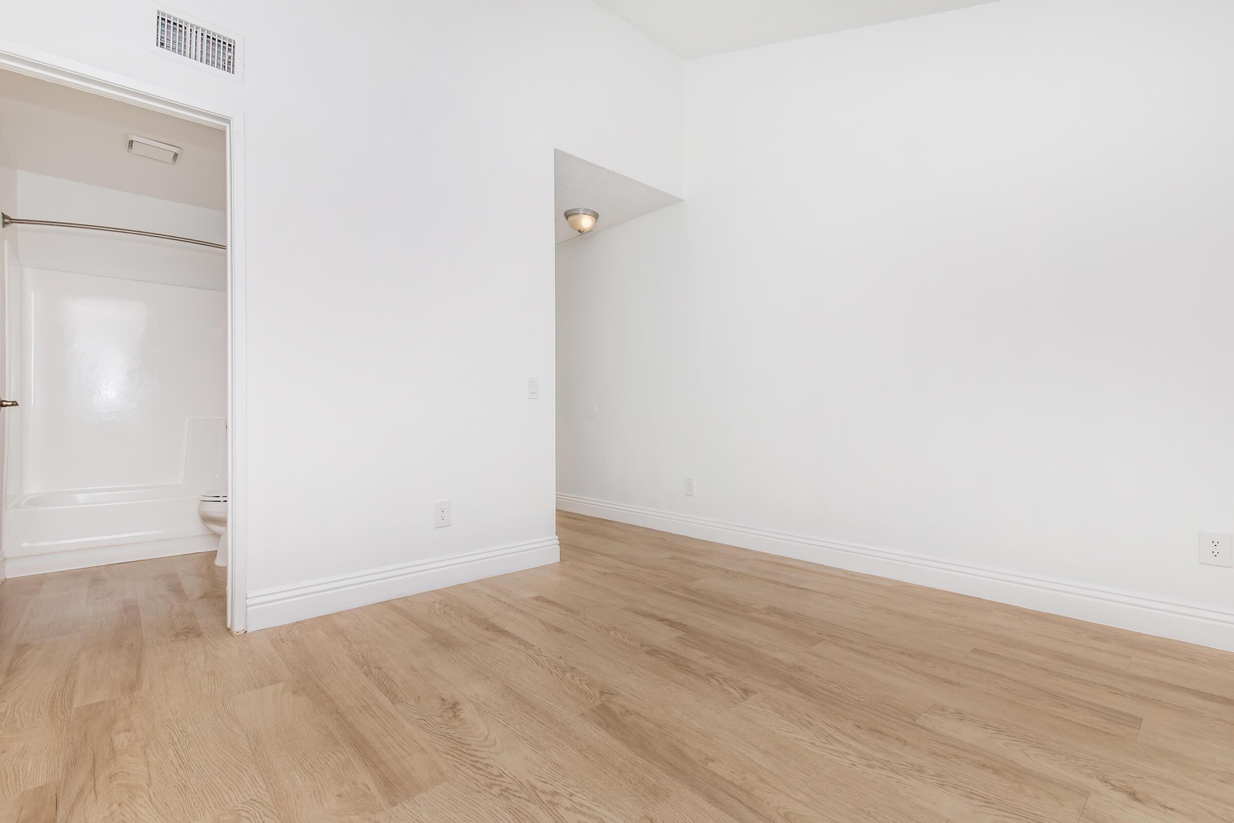 A spacious, empty room with light-colored wood flooring. There's a doorway leading to a bathroom area with a shower and toilet visible. The walls are painted white, and the ceiling is high, allowing for ample natural light. The overall ambiance is bright and minimalistic.