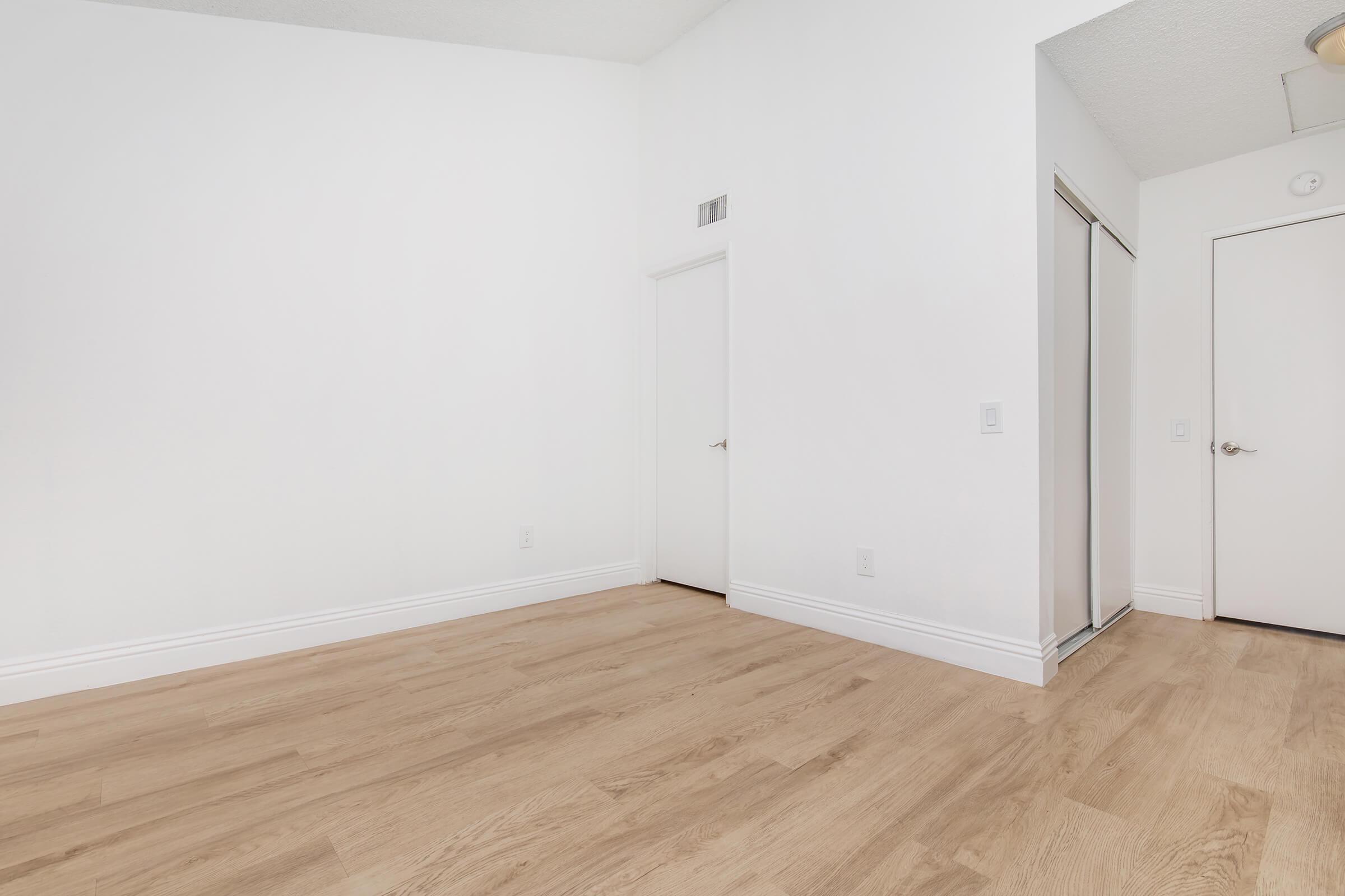 Empty room with light-colored wooden flooring and white walls. There are two doors: one leading to a closet and another that appears to lead outside the room. Natural lighting creates a bright and spacious feel. The room is minimalist with no furniture or decorations.