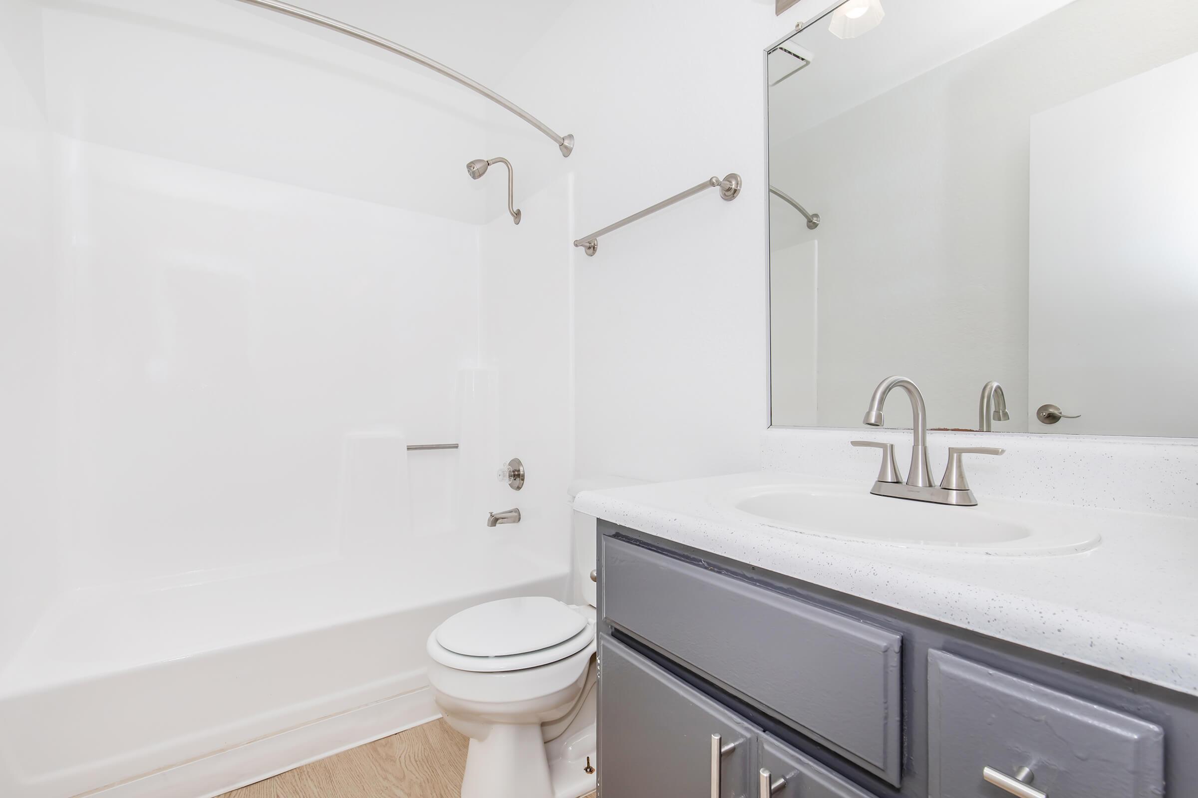 A clean, modern bathroom featuring a white bathtub with a curved shower rod, a sink with a silver faucet, and a large mirror above the sink. The walls are painted white, and there are gray cabinets under the sink. The flooring is light-colored wood.