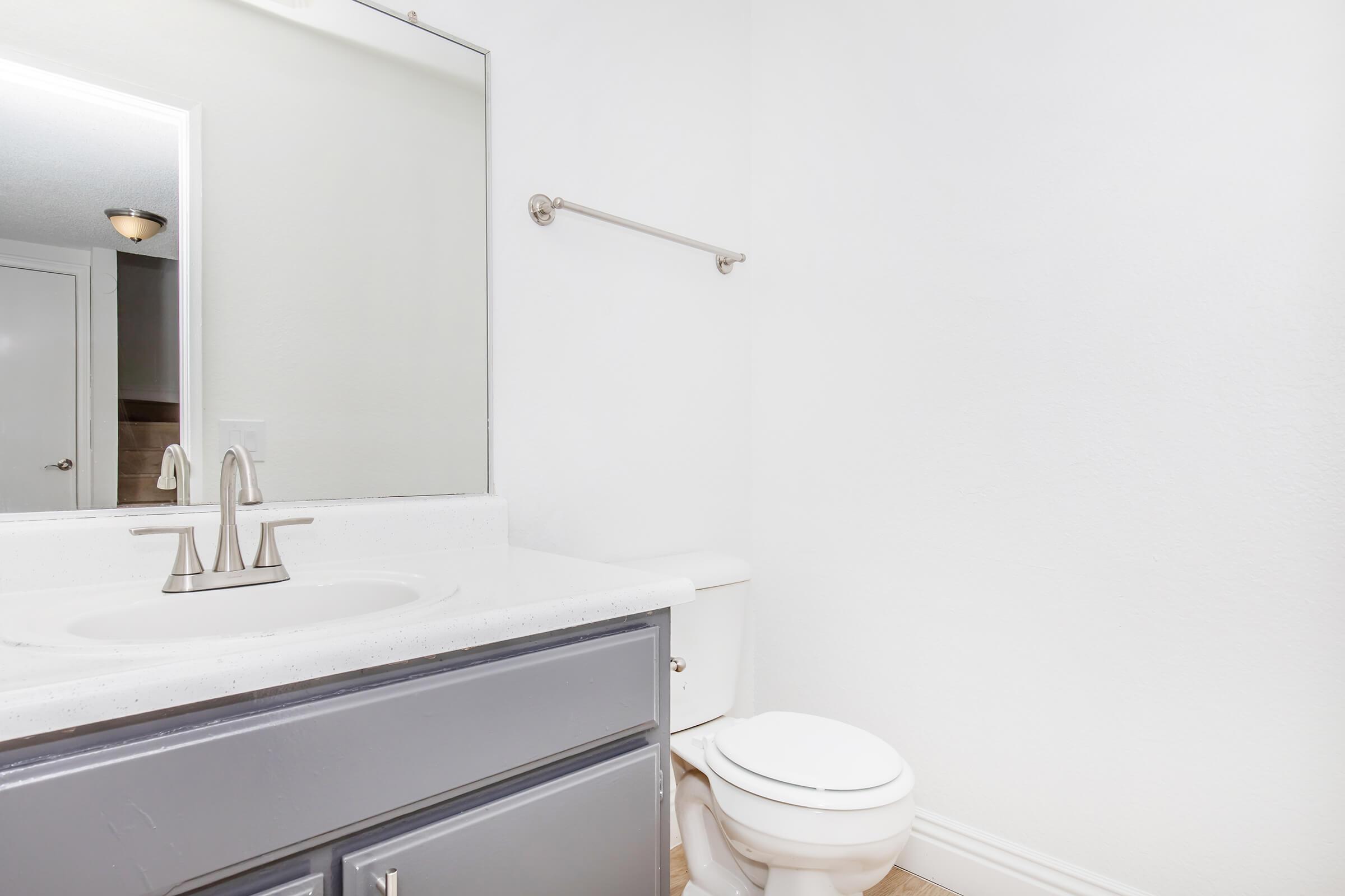 A clean, modern bathroom featuring a gray vanity with a white countertop and sink, a large mirror above the sink, and a white toilet. The walls are painted white, and the flooring is light-colored. A towel bar is mounted above the vanity. The overall look is bright and minimalist.