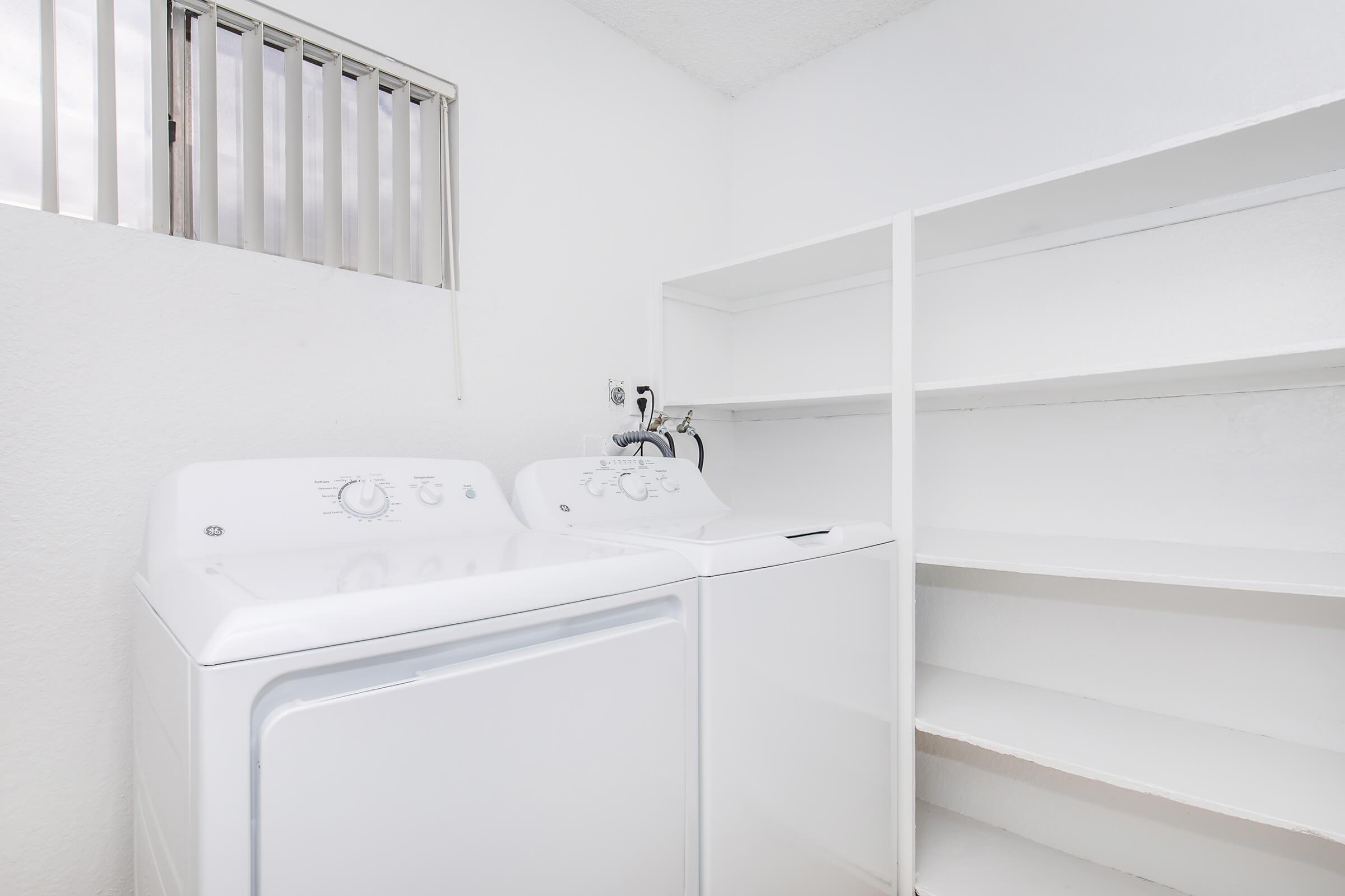 A laundry room featuring a white washing machine and dryer side by side, with a small window above them. There are empty white shelves on the wall next to the appliances, and the walls and floor are painted white, creating a bright and clean atmosphere.