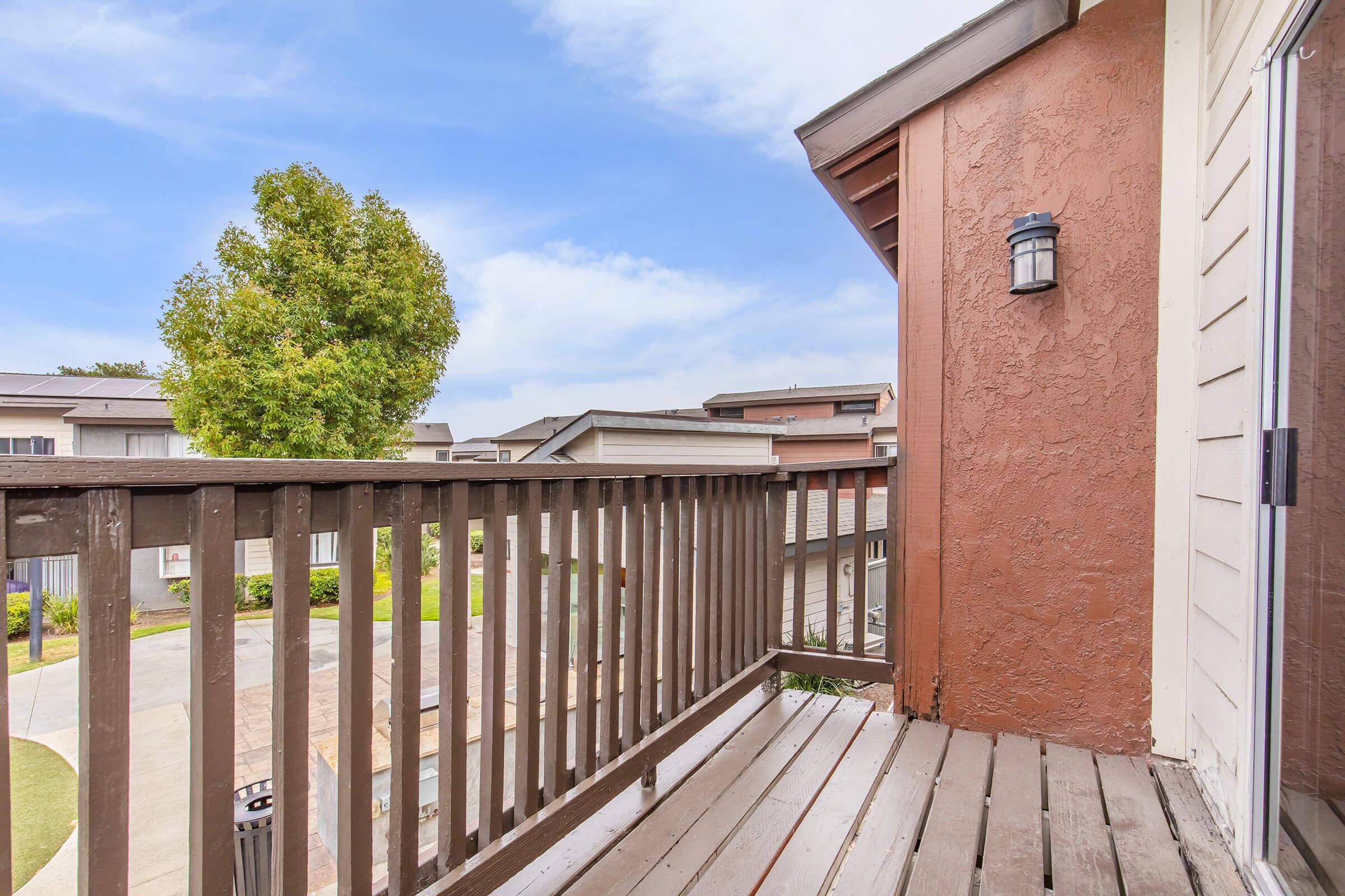 A view from a balcony overlooking a residential area. The wooden railing frames the scene, and a green tree is visible in the background against a blue sky with scattered clouds. The color of the building is a warm terracotta, adding to the inviting atmosphere of the space.