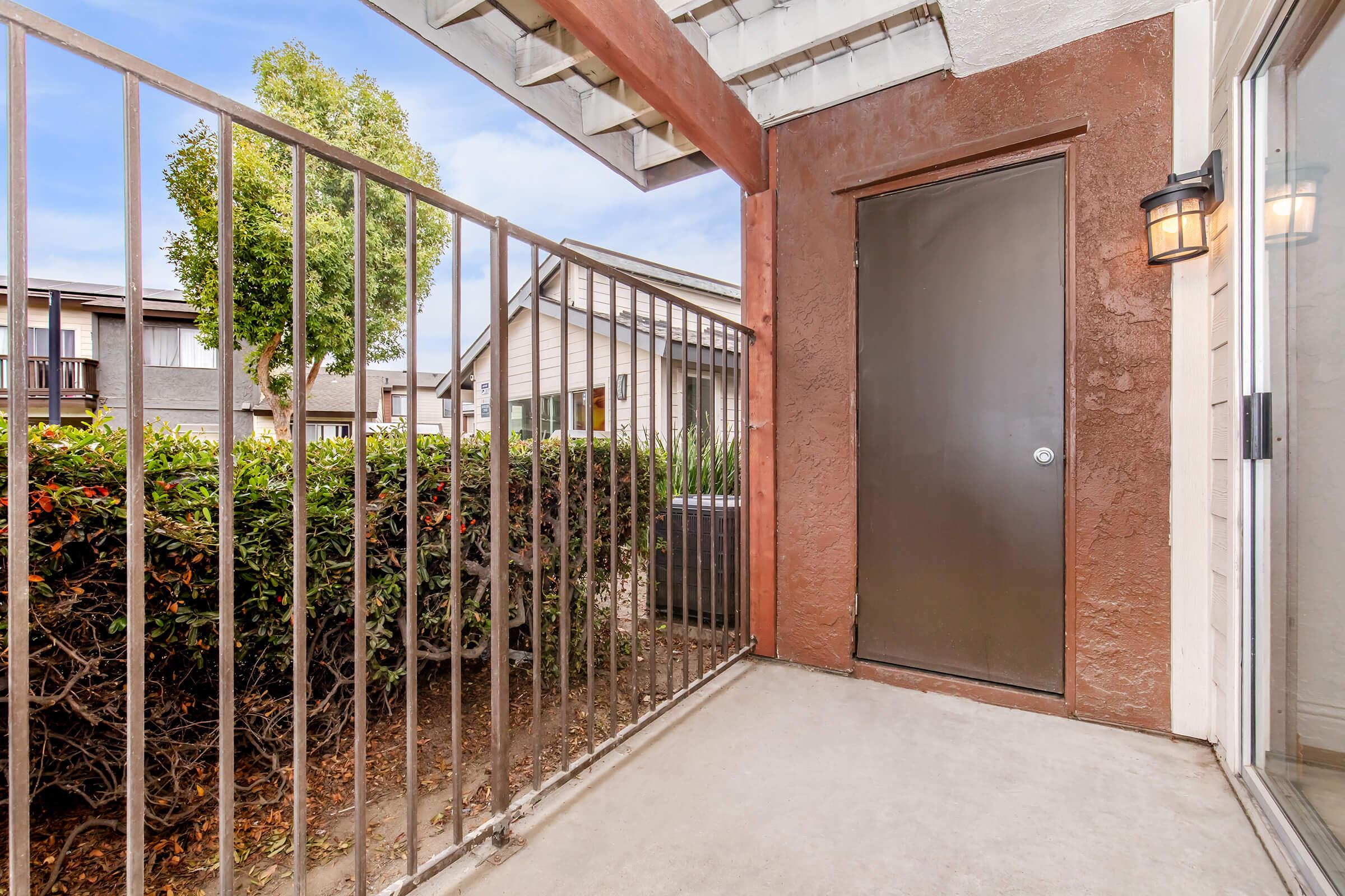 A view of a concrete patio area with a metal railing, bordered by a lush green hedge. A brown door is visible on the right, and beyond the railing, there are buildings in the background under a clear blue sky. The scene suggests a residential outdoor space.
