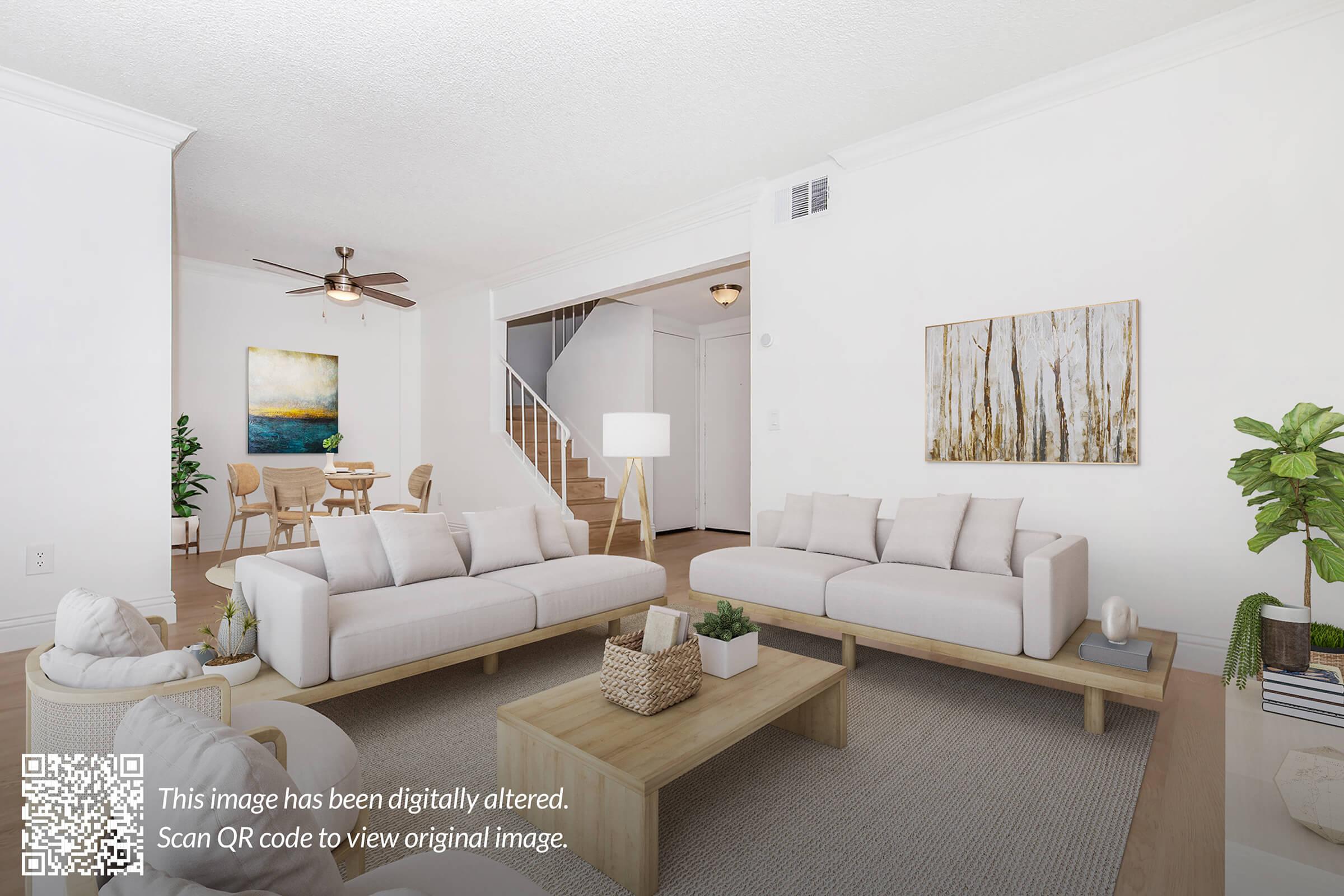 A modern living room featuring two light-colored sofas, a wooden coffee table, and a neutral rug. A staircase is visible in the background, along with a decorative lamp and a piece of wall art depicting trees. The walls are painted white, creating a bright and airy atmosphere.