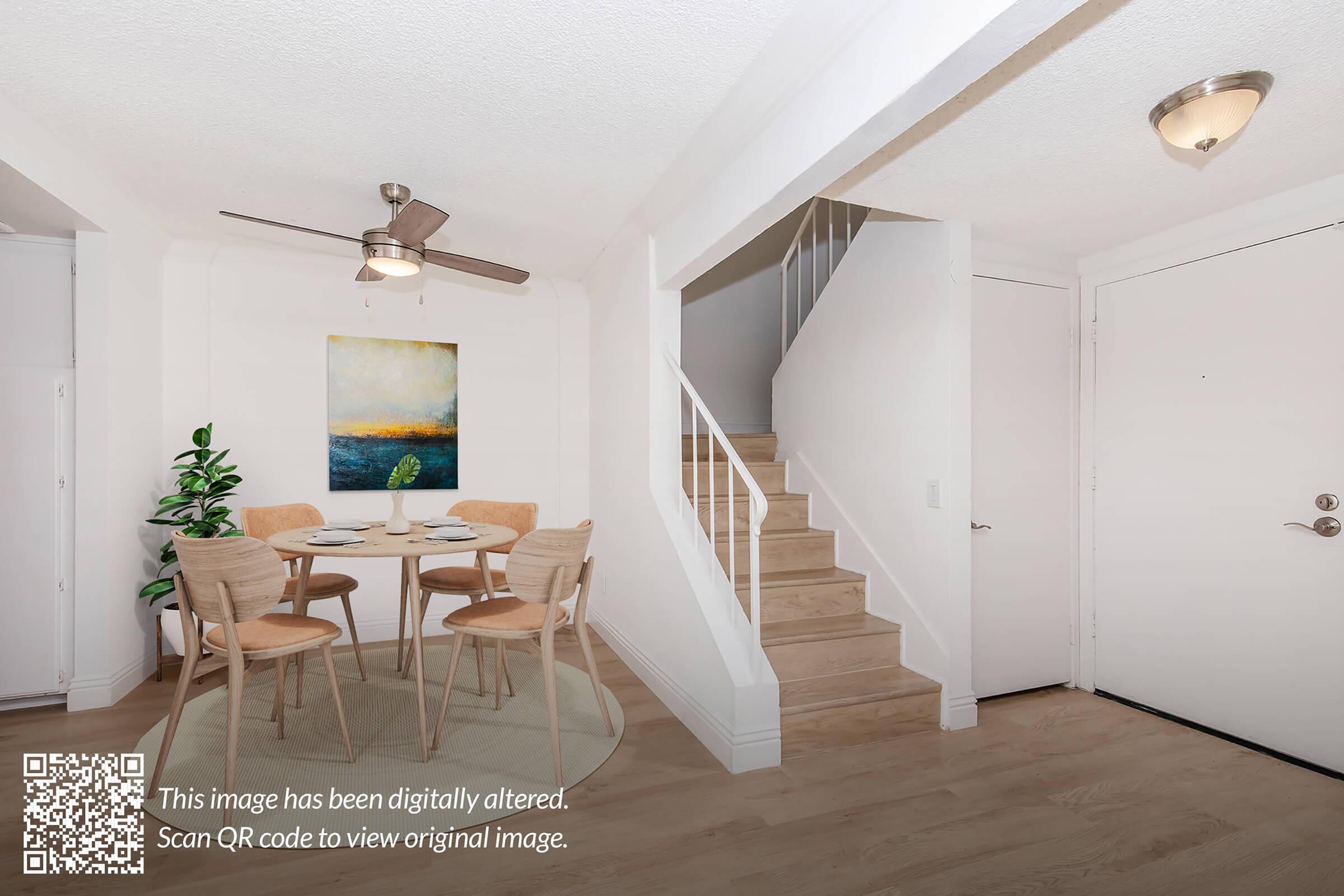 A bright, modern interior with a dining area featuring a round table and four wooden chairs. A colorful abstract painting hangs on the wall. Stairs lead to an upper level, and a front door is visible. The room has light-colored walls and a ceiling fan, creating a spacious, inviting atmosphere.