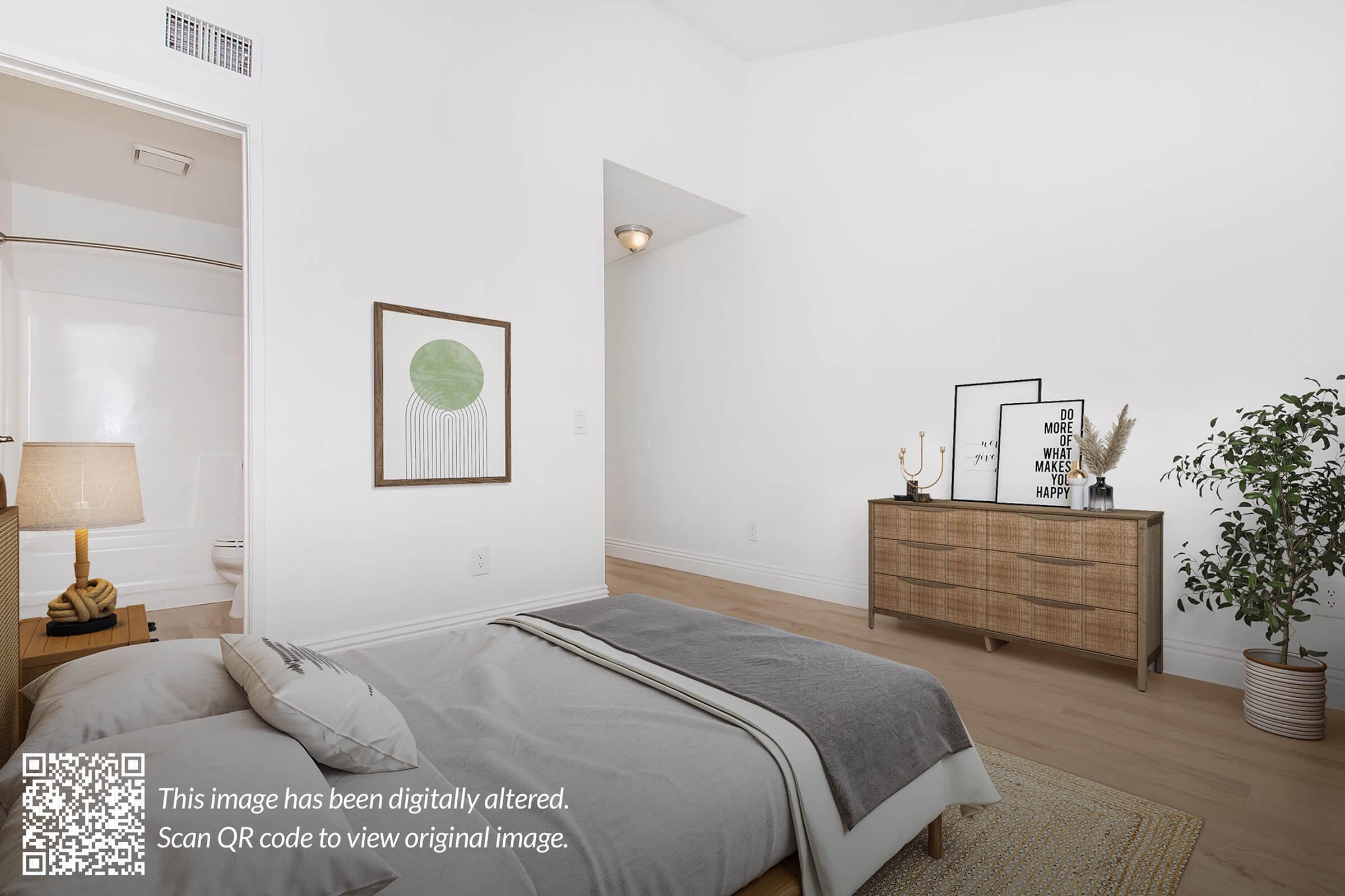 A serene bedroom with a light-colored theme, featuring a neatly made bed with a gray duvet. A wooden dresser with decorative items stands against the wall, and a potted plant adds a touch of greenery. A framed artwork hangs above the bed, and there is a doorway leading to a bathroom in the background.